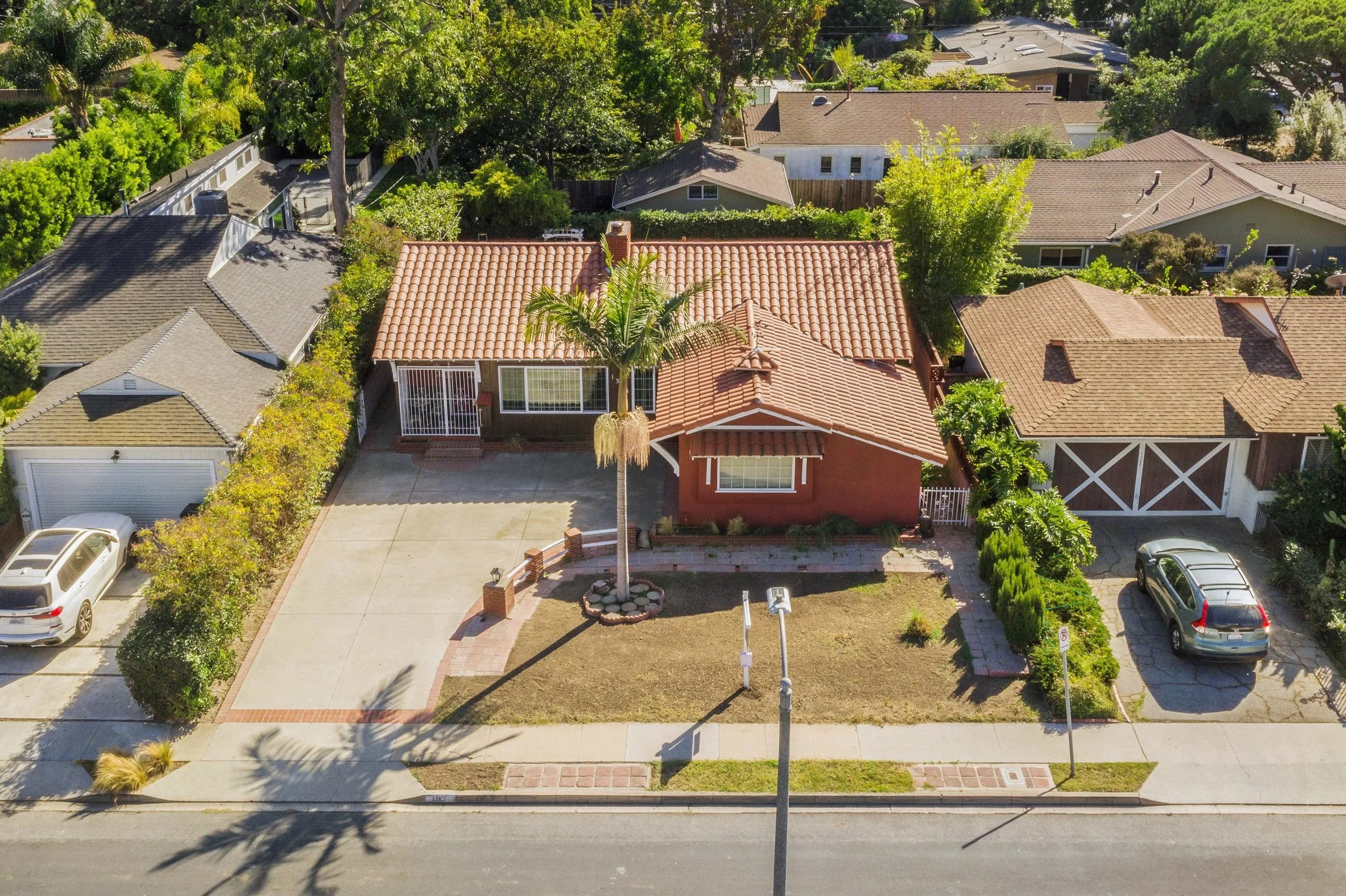 Aerial view of a residential neighborhood featuring several houses with various roof styles and colors, a large landscaped front yard with a palm tree, and parked cars on the street.