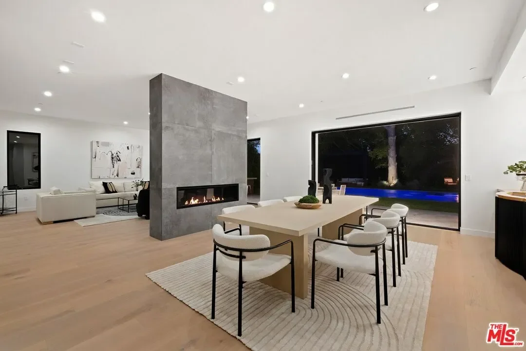 Modern open-concept living and dining room with a large fireplace, white and black furniture, wooden floors, and a wall-sized window showing outdoor trees at night.