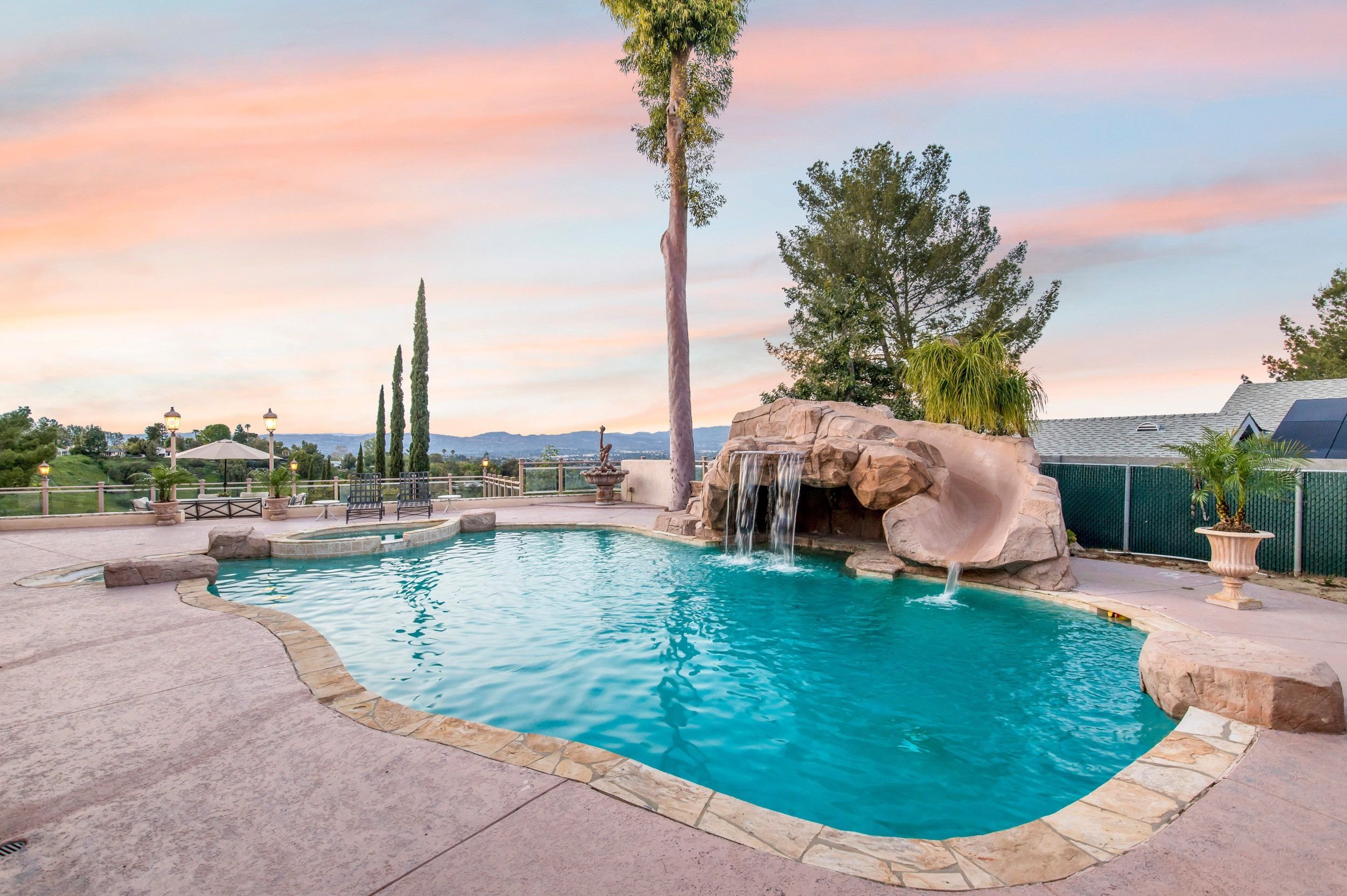 A backyard swimming pool with a waterfall feature, surrounded by rocks and potted plants. There are tall trees and a mountain view in the background, with a pastel-colored sky at sunset or sunrise.