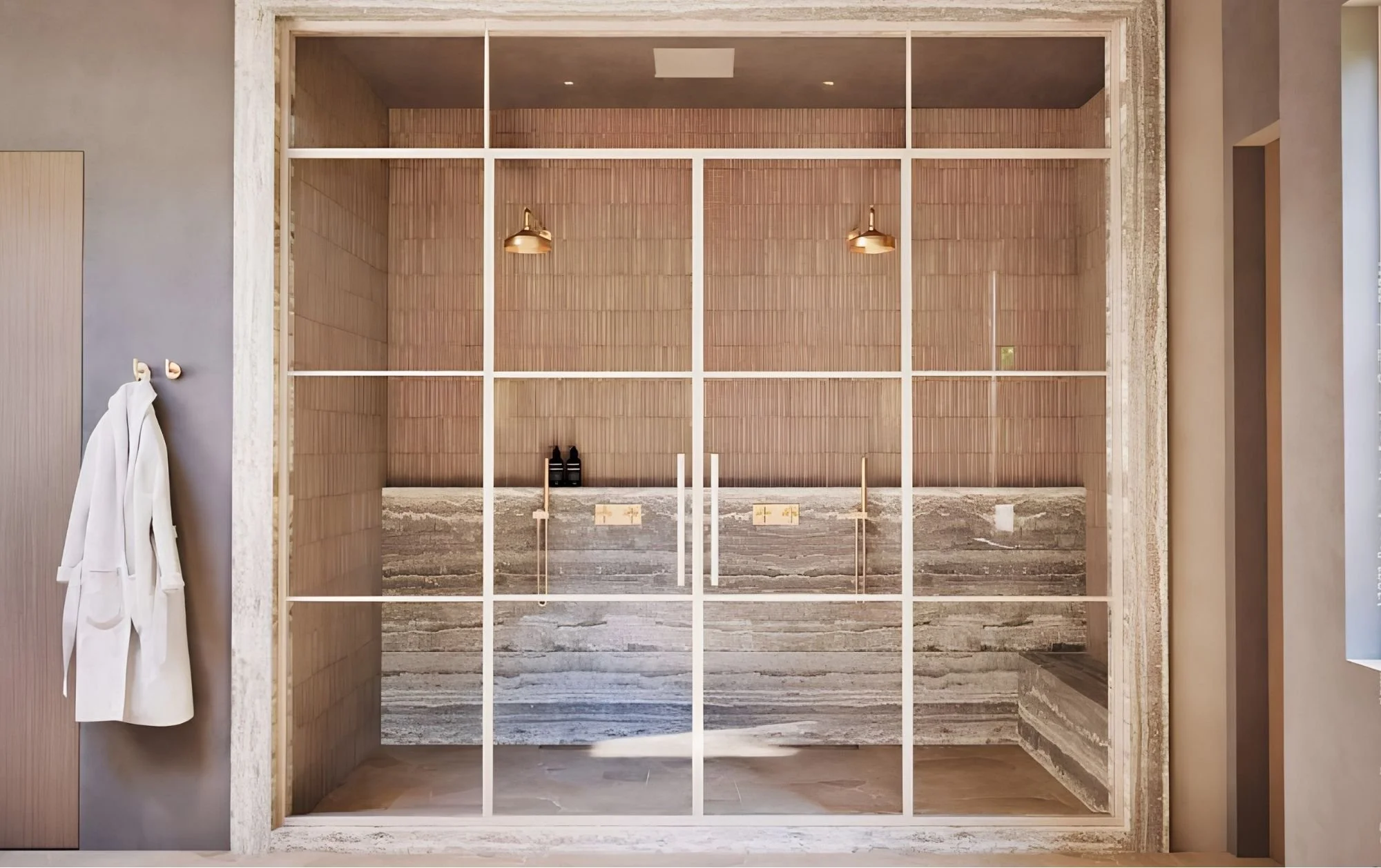 Interior view of a modern bathroom under construction, with a large glass window or door opening framed by white wood, showing a vanity area with a granite countertop and pipes below, and two brass light fixtures hanging from a wooden-paneled wall in
