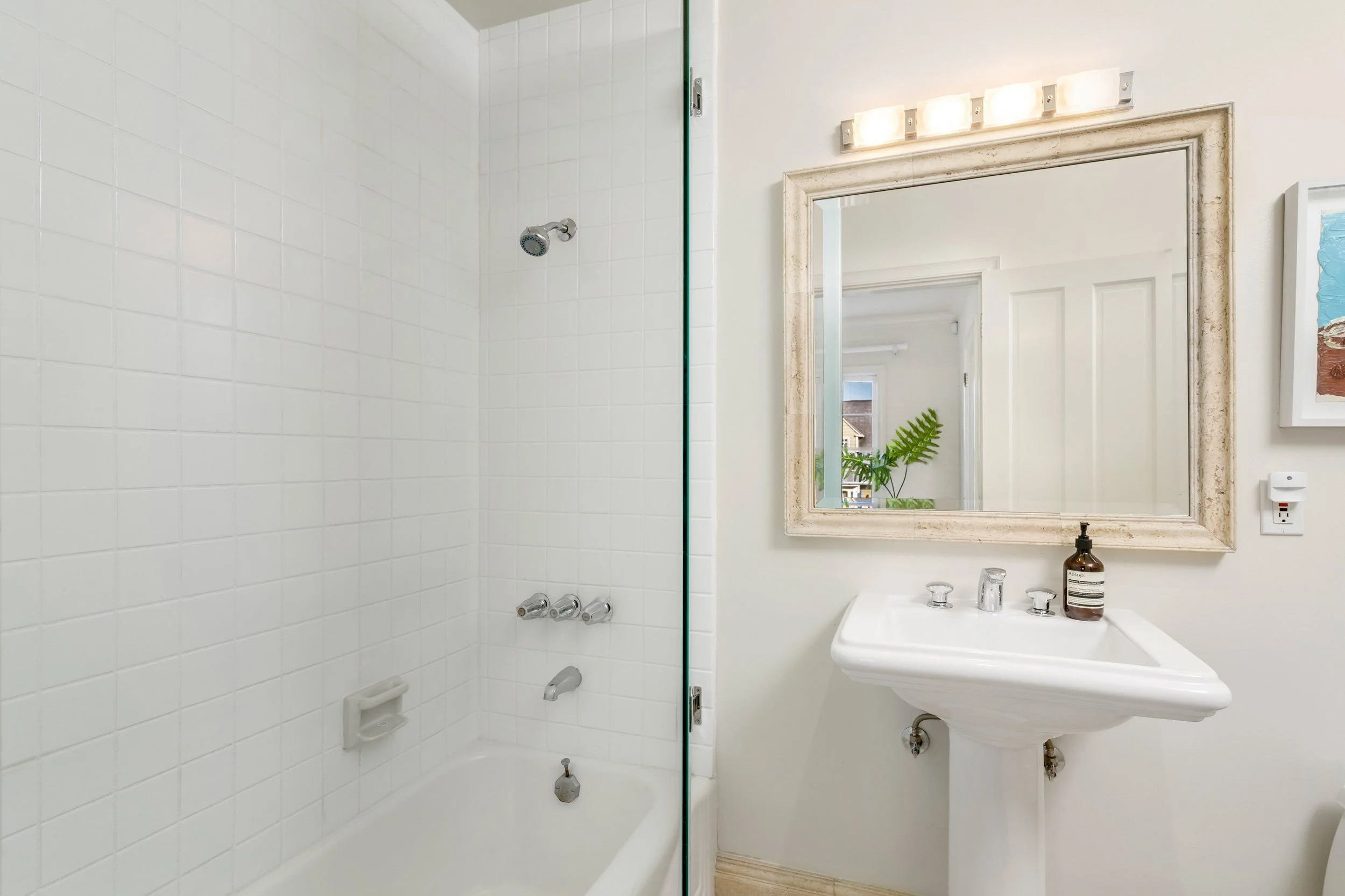 Bathroom with a shower on the left and a sink with a mirror above it on the right. The shower has white tiles and a showerhead. The mirror has a beige frame. There's a window with a view of rooftops and trees outside. A plant is visible through the m