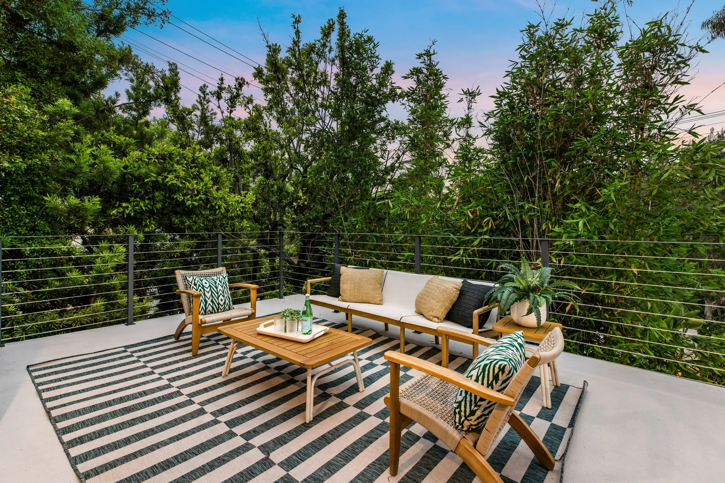 Outdoor patio with seating area, including a sofa, armchairs, and coffee table, decorated with patterned cushions and a striped rug, surrounded by lush green trees and plants, with a sky showing sunset colors.