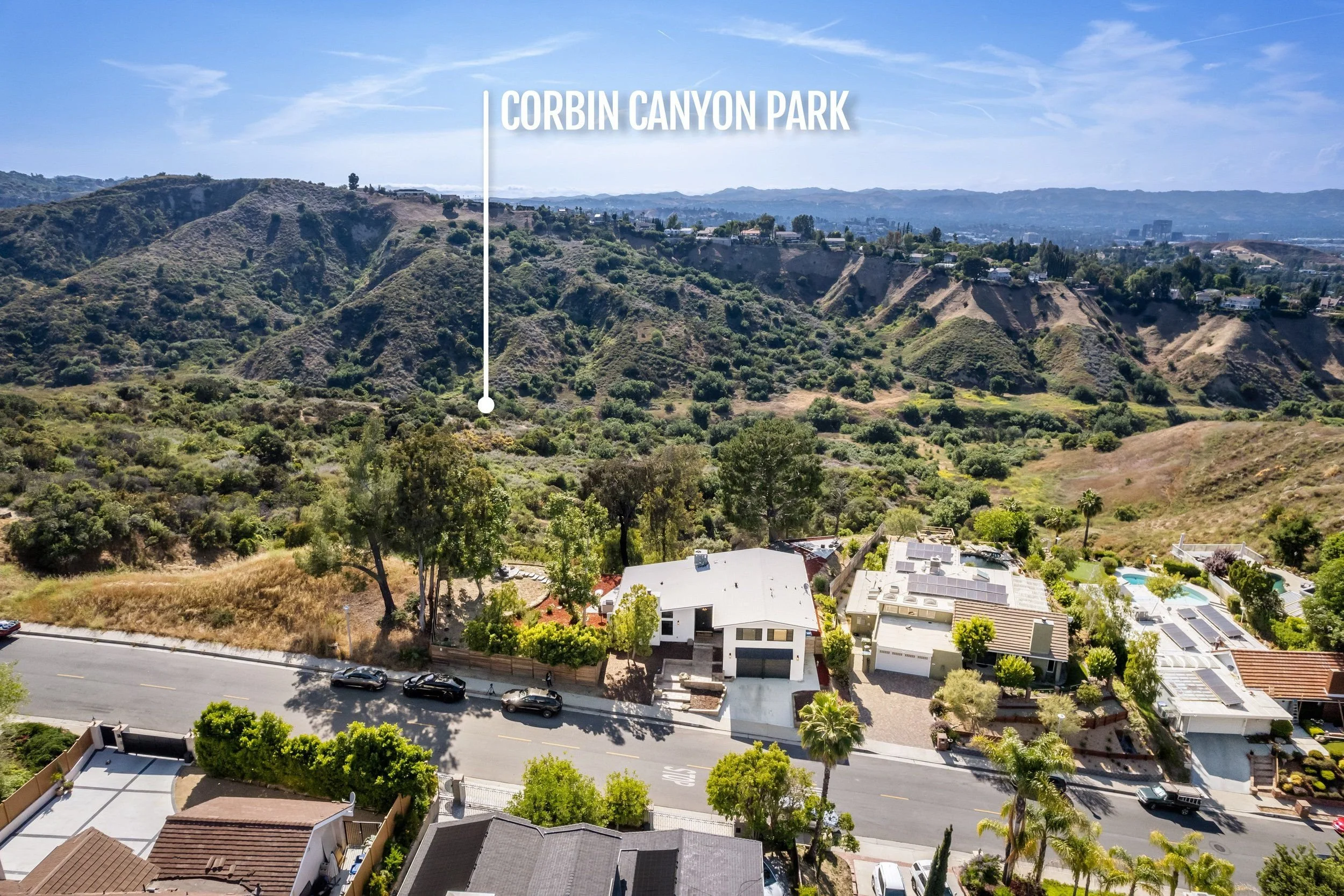 Aerial view of Corbin Canyon Park showing residential houses, a street with parked cars, a hillside with trees and bushes, and distant mountains under a partly cloudy sky.