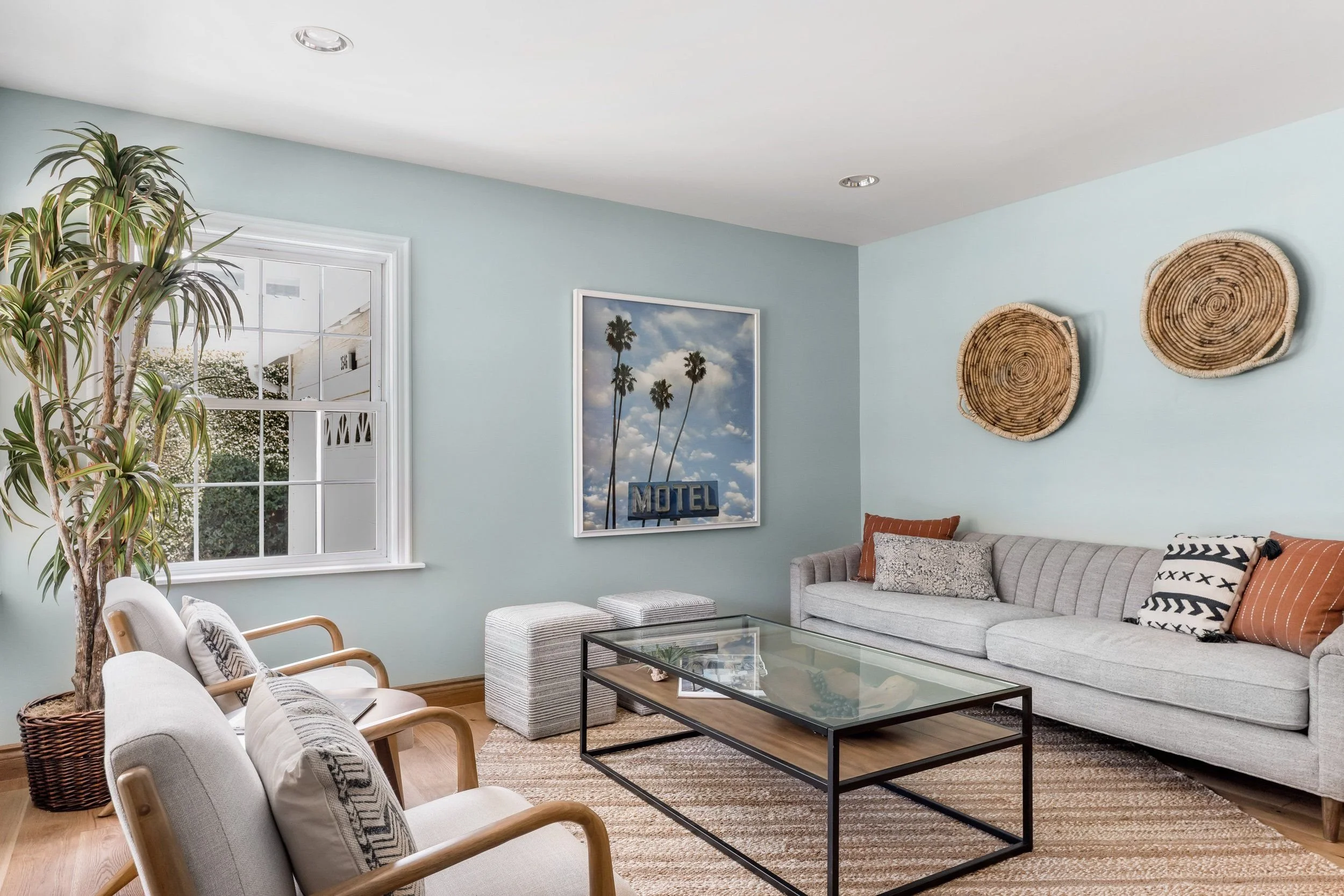 Living room with light blue walls, a large window with white trim, decorated with houseplants, framed poster of palm trees with 'MOTEL' text, wicker wall art, a gray sofa with patterned throw pillows, a glass coffee table, and a beige armchair with a