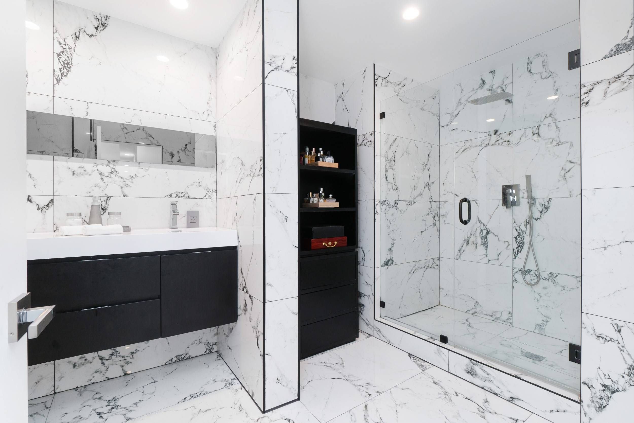 A modern bathroom featuring white marble tiles with black veining on the walls and floor, a black vanity with a white countertop, and a large mirror. There is a tall black shelving unit with various toiletries and a glass shower enclosure with a rain