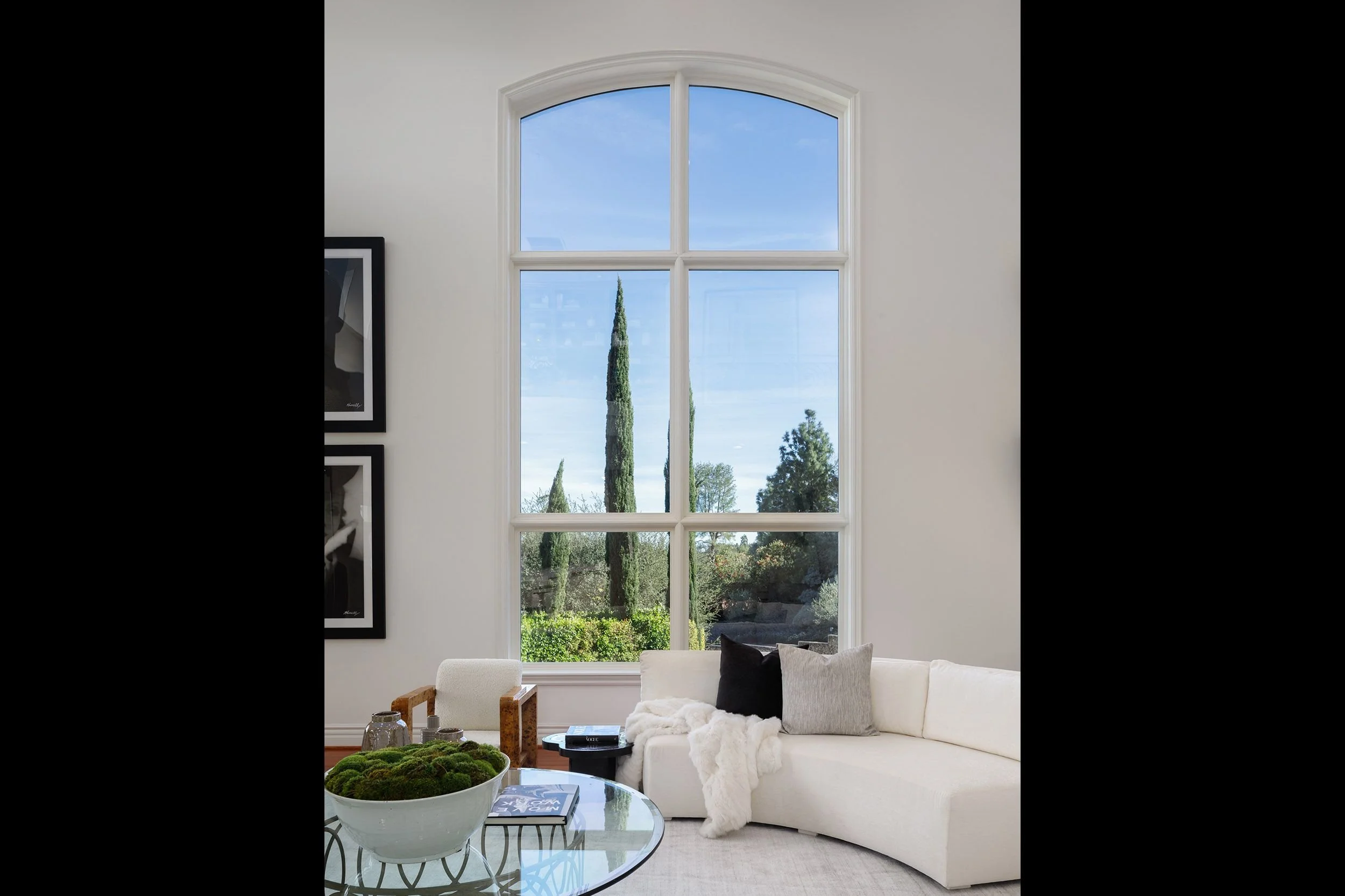 Living room with large window, white sofa with pillows and throw blanket, glass coffee table with greenery, and framed artwork on the wall.
