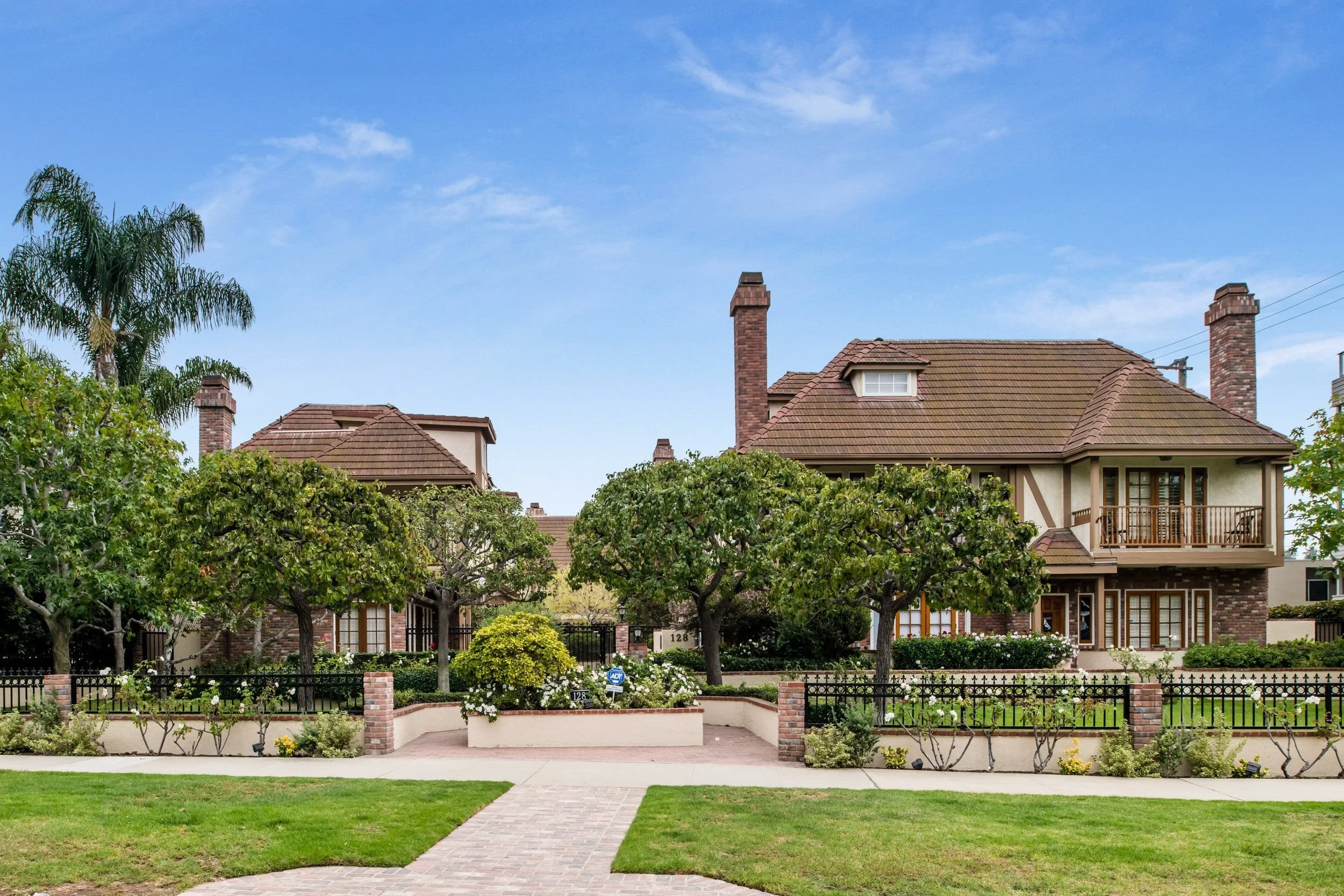 A view of large residential house with multiple chimneys, surrounded by trees and a well-maintained yard with a brick pathway and a small garden wall.