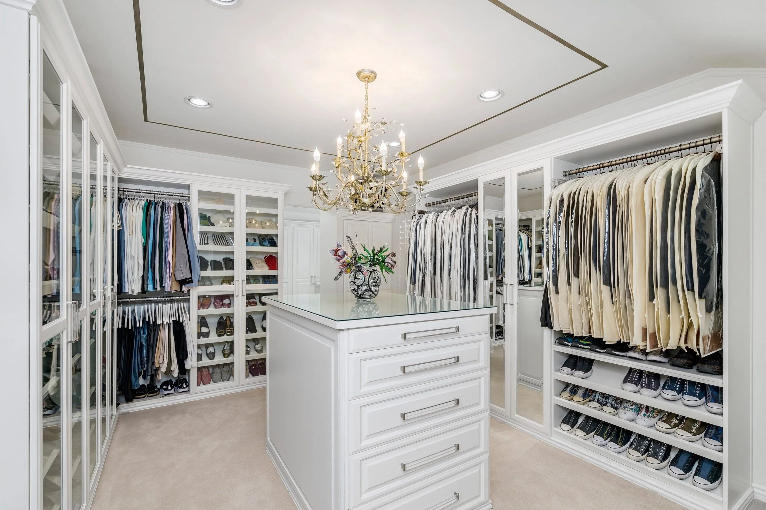 A walk-in closet with white built-in shelves filled with clothes, shoes, and accessories. There is a white island with drawers in the center, a chandelier hanging from the ceiling, and a mirror on the left wall.