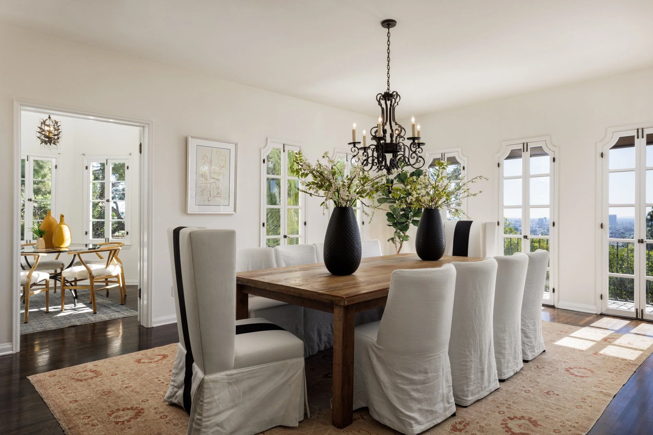 A dining room with a large wooden table surrounded by white slipcovered chairs, a black chandelier hanging above, and large windows with views of greenery and cityscape outside. Two black vases with floral arrangements sit on the table.