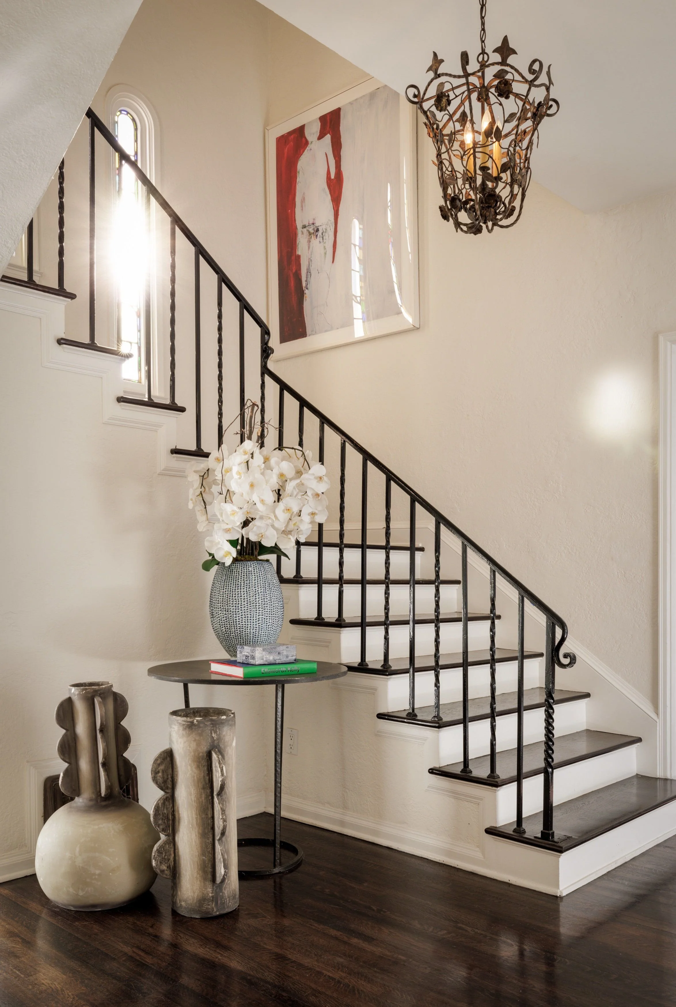 Interior view of a staircase area with a black metal railing, a large abstract portrait painting on the wall, white walls, and a decorative chandelier hanging from the ceiling. A small round table with a vase of white orchids and books is in the fore