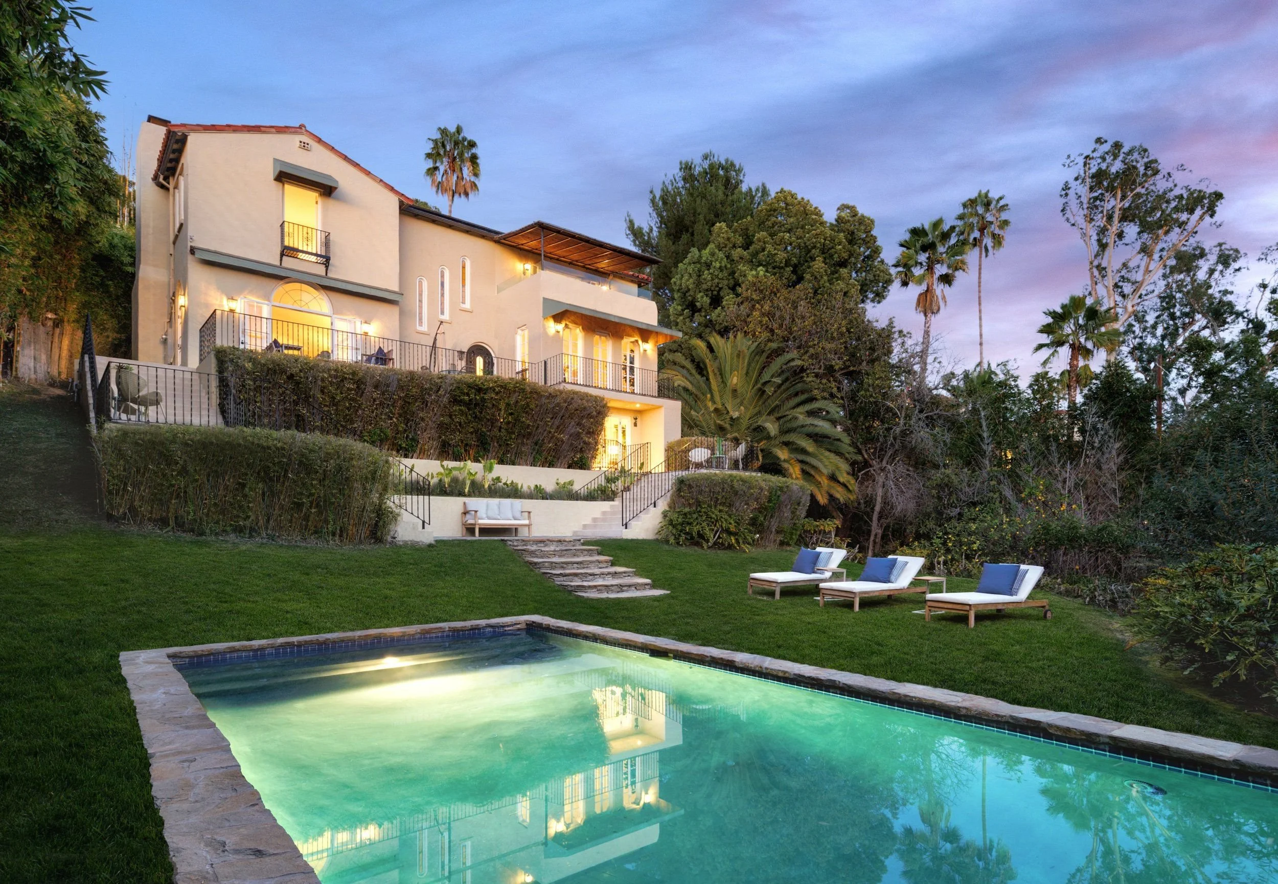 A large multi-story house with beige walls and red tile roof, surrounded by lush greenery and tall palm trees, with outdoor lighting and balconies. In the foreground, there is a fenced swimming pool with a stone deck, outdoor furniture, and well-main