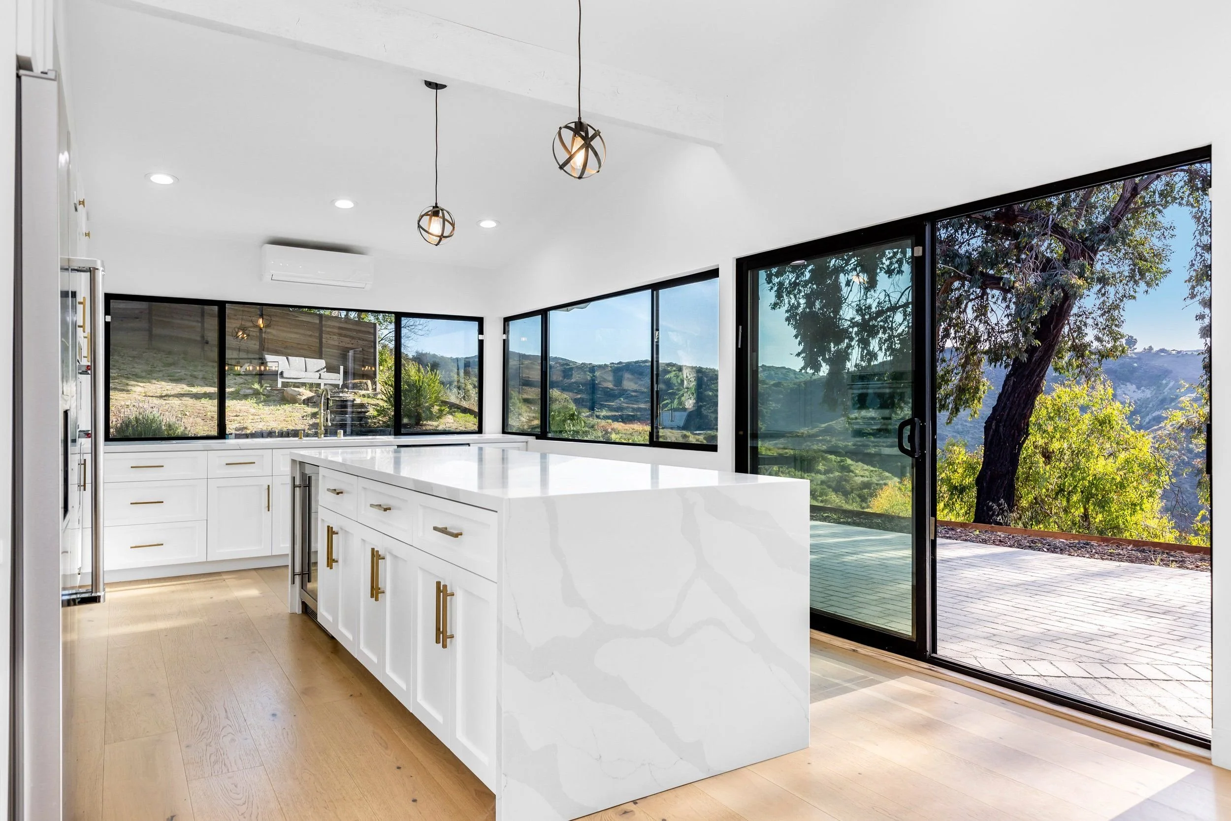 Modern kitchen with white cabinets, large white island, wooden floor, and large sliding glass doors revealing outdoor greenery and hills.