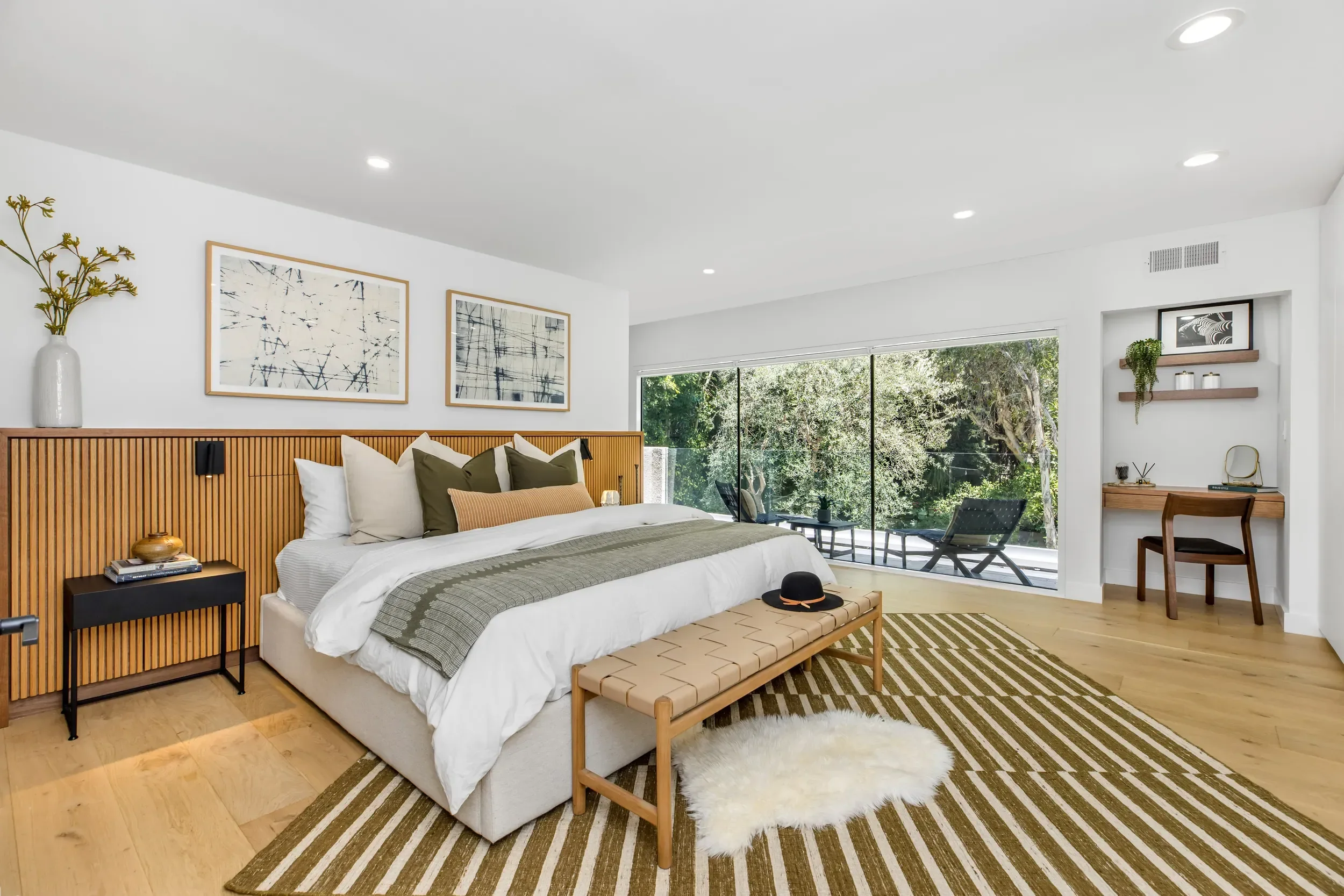 A modern bedroom with a large bed, decorative pillows, a wooden bench, a black side table, and a white shaggy rug. There are large windows showing trees outside, and artwork and shelves with decorative items on the walls.