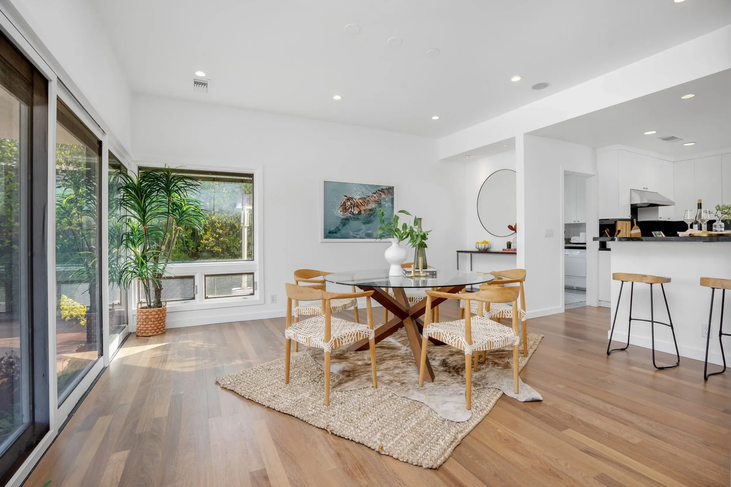 Modern dining room with wooden table, six chairs, a woven rug, large windows with view of greenery, white walls, hanging artwork, and kitchen area with white cabinetry and black countertop.