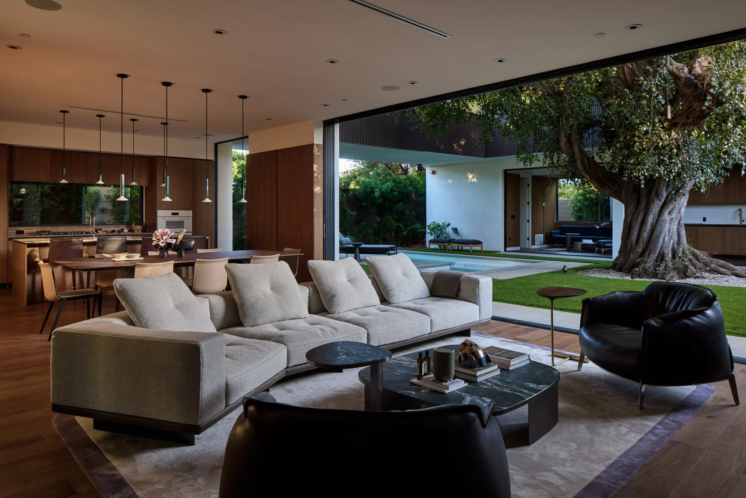 Modern open-plan living room with large glass doors opening to a backyard garden with a tree and outdoor seating. Inside features a beige sectional sofa, black and beige chairs, a round coffee table, and a dining area with hanging pendant lights. Woo