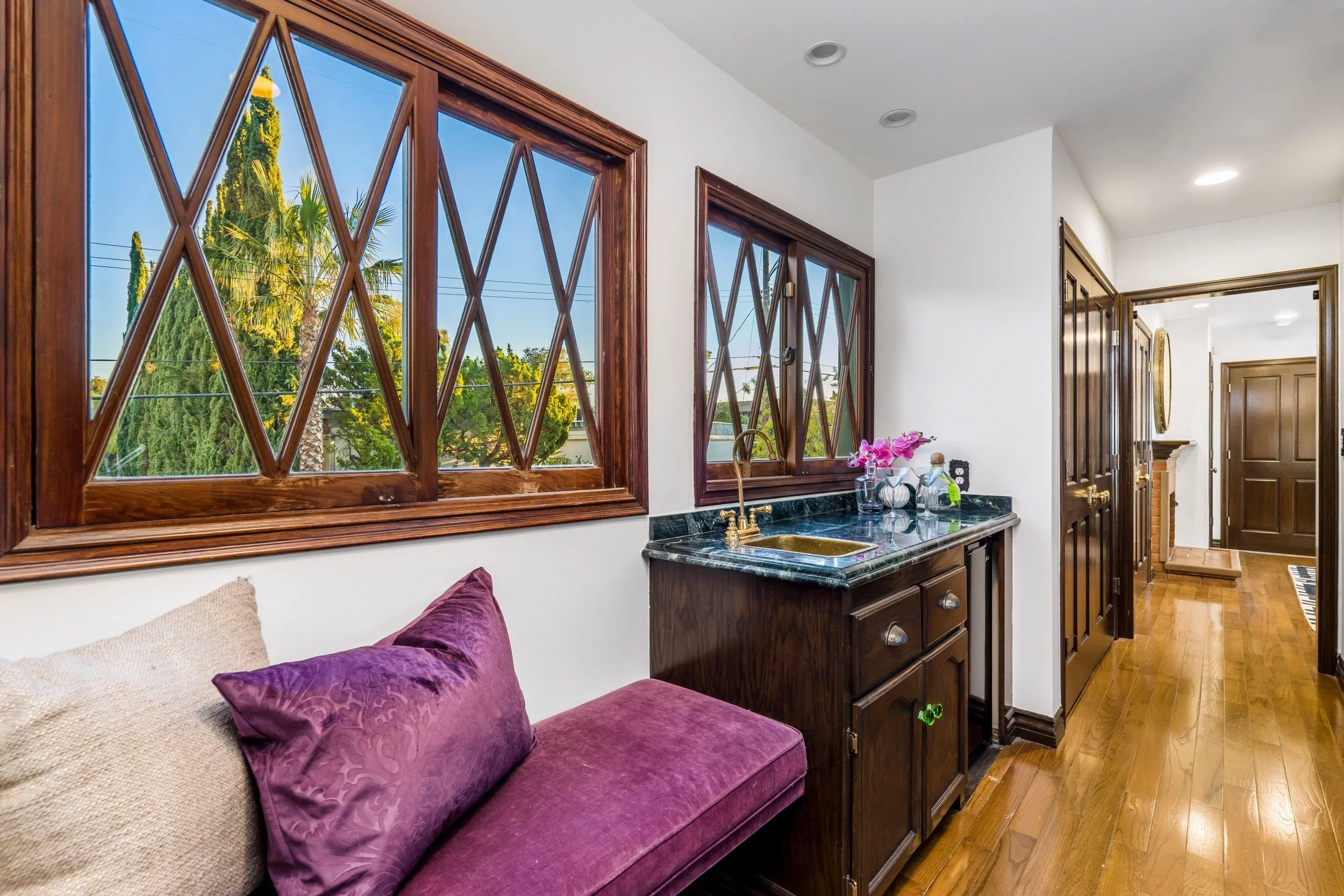 View from inside a kitchen with tropical landscape visible through diamond-pane wooden windows. Includes maroon and beige cushions on a purple upholstered bench, a black granite counter with a bronze sink, pink flowers in a glass vase, and wooden cab
