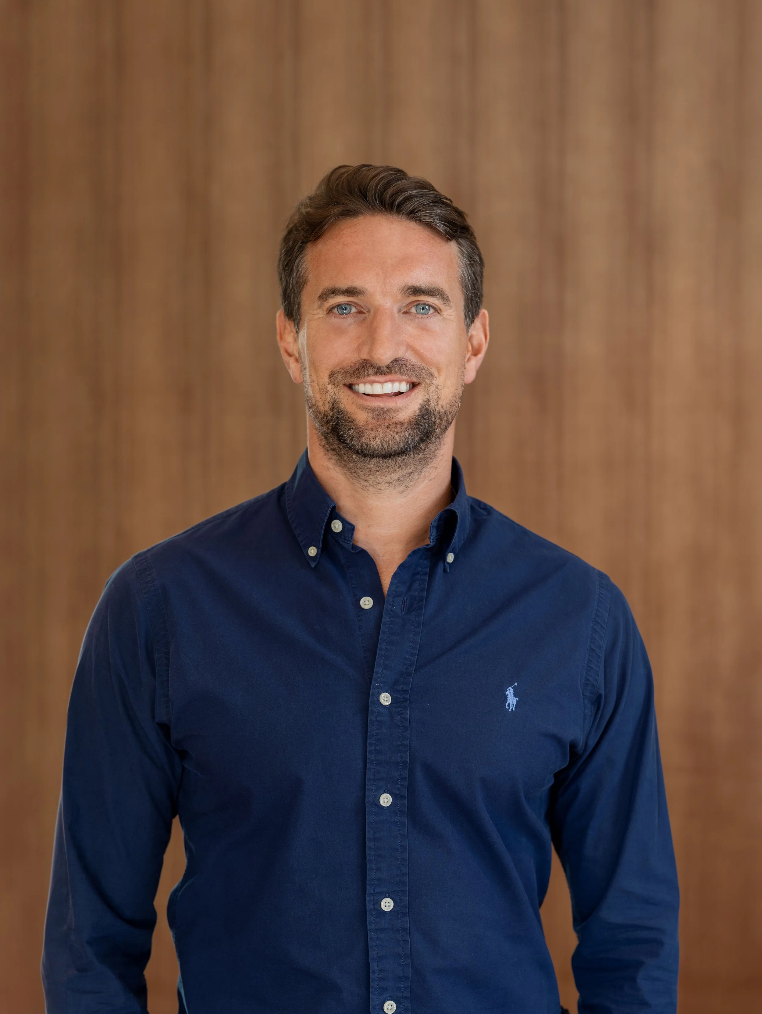 A smiling man with blue eyes, brown hair, and a beard, wearing a navy blue button-up shirt with a small logo on the chest, standing against a wooden background.