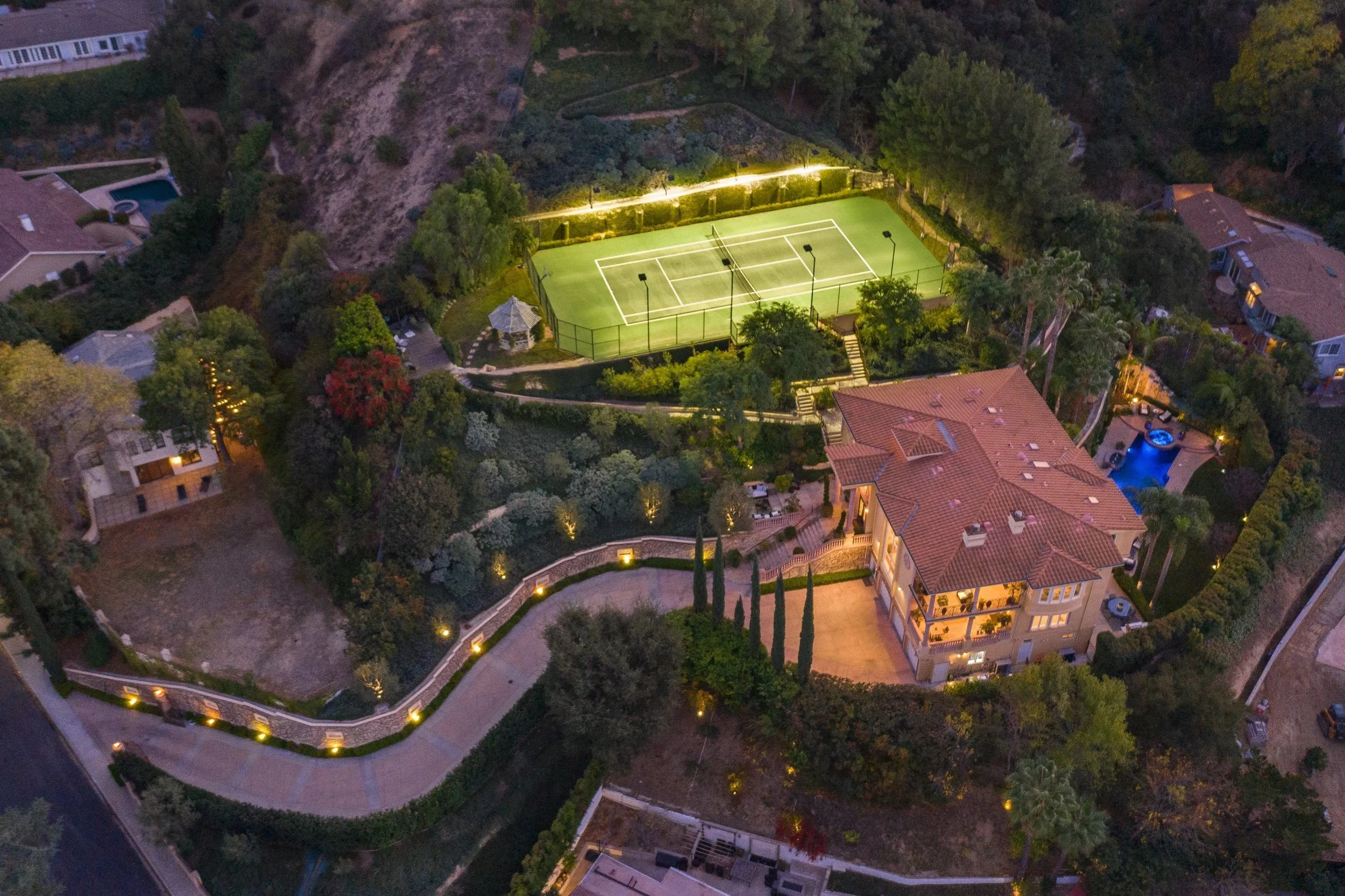 Aerial night view of a residential neighborhood with a tennis court illuminated by lights, surrounded by trees and houses.