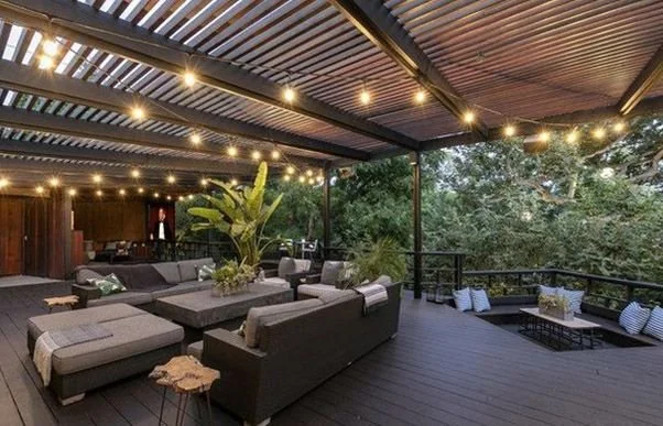 Outdoor rooftop patio with black sofas, potted plants, string lights, and surrounding trees.