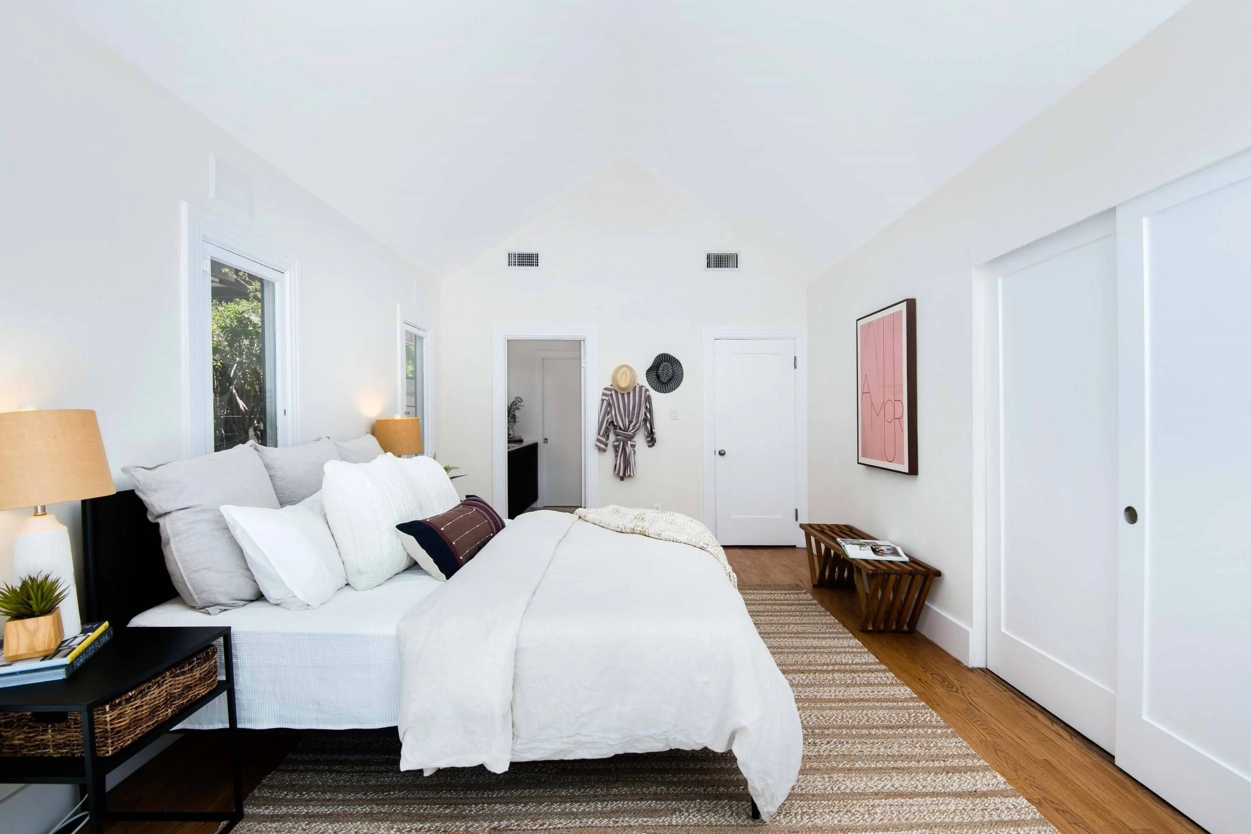 A cozy bedroom with a white bed, beige bedside lamps, two windows, wall art, and a wooden floor.