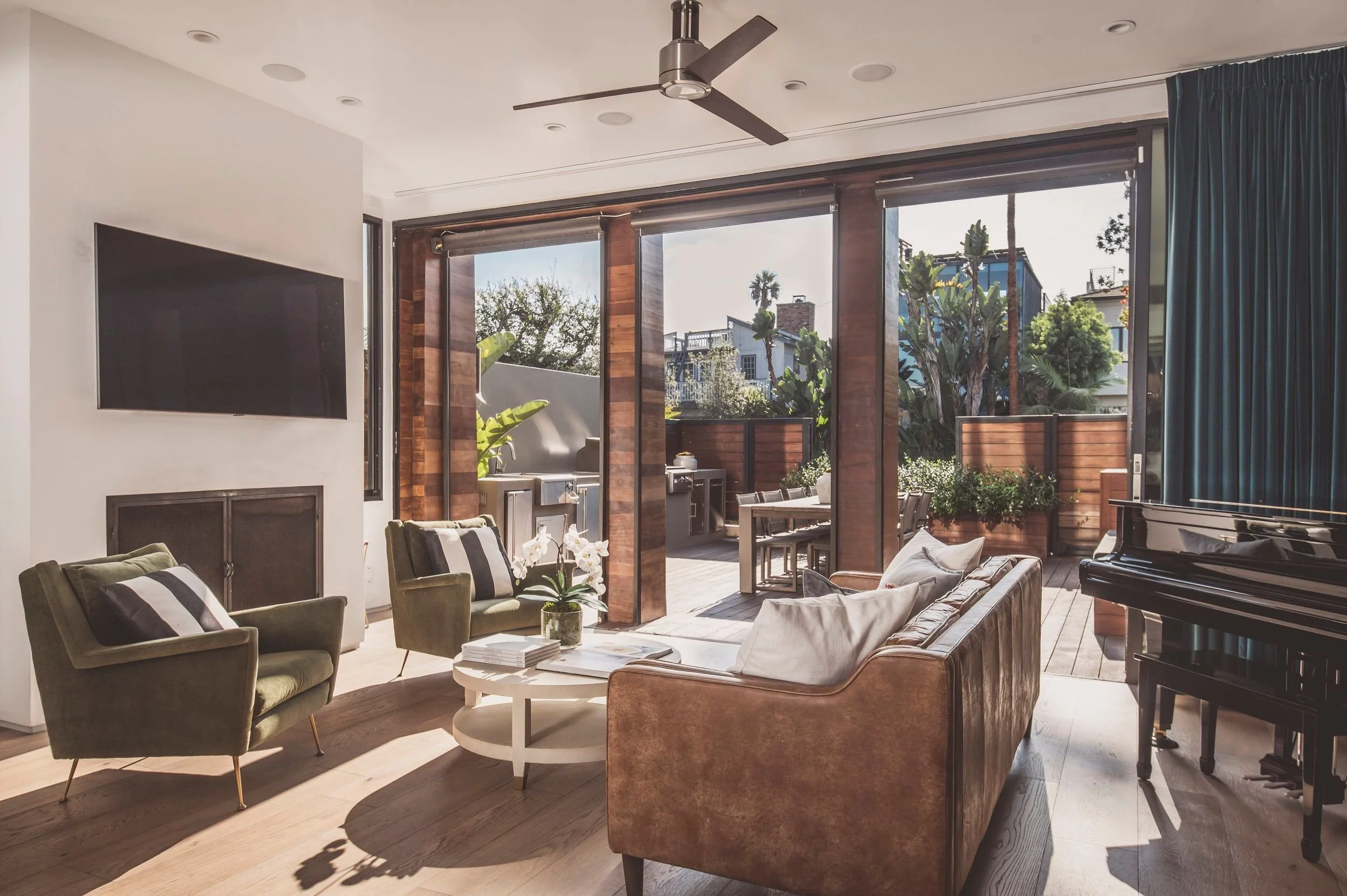 Living room with green armchairs, a brown leather sofa, a black grand piano, and large glass doors opening to an outdoor patio with greenery and trees.