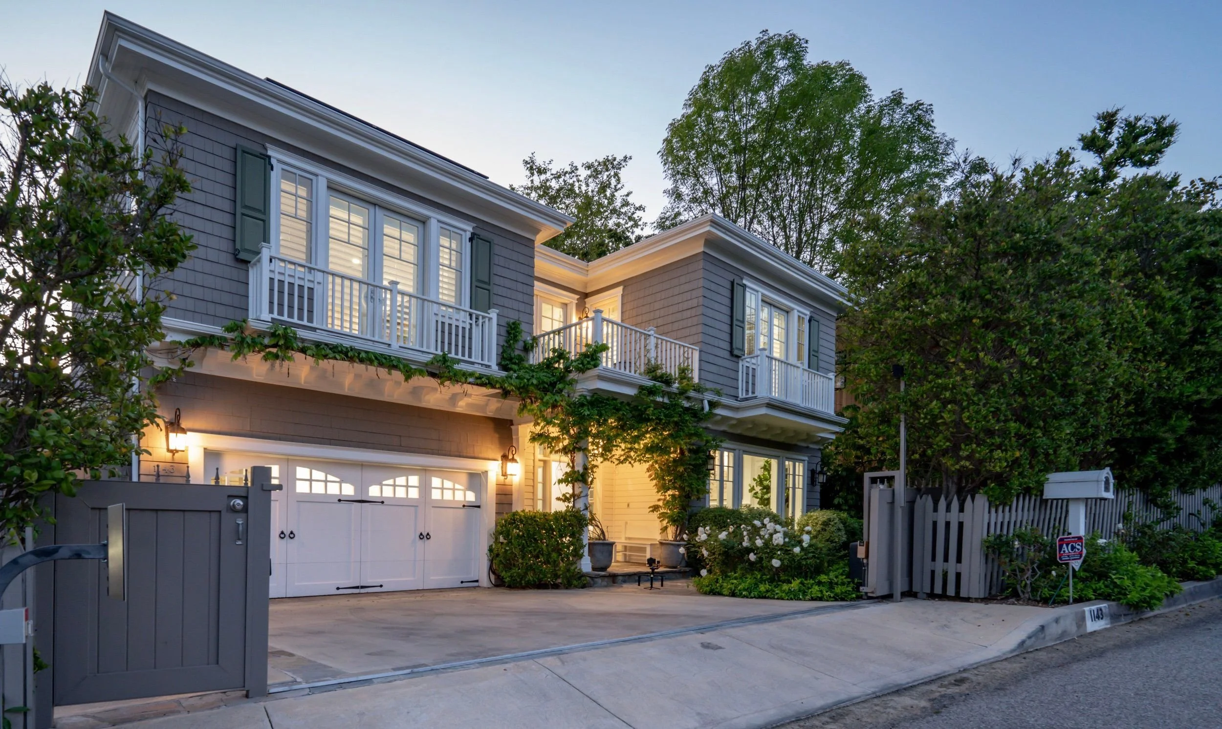 Two-story house with gray siding, white trim, and green shutters. Front yard with trees, shrubs, and a white fence, illuminated by outdoor lights at dusk.