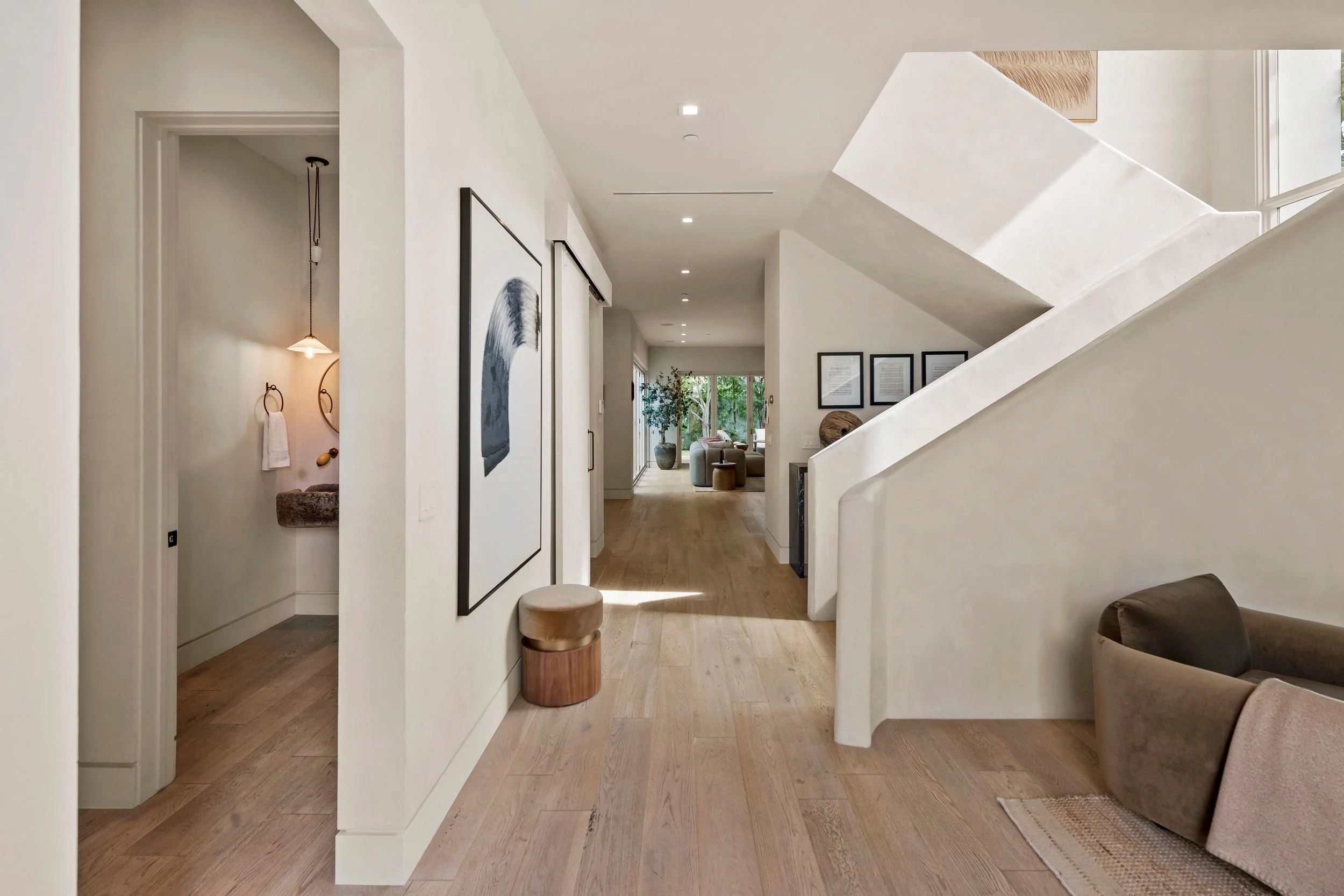 A modern interior home entryway with art on the walls, wooden floors, a small stool, a staircase with white walls, and a view of a living room with window and outdoor plants.