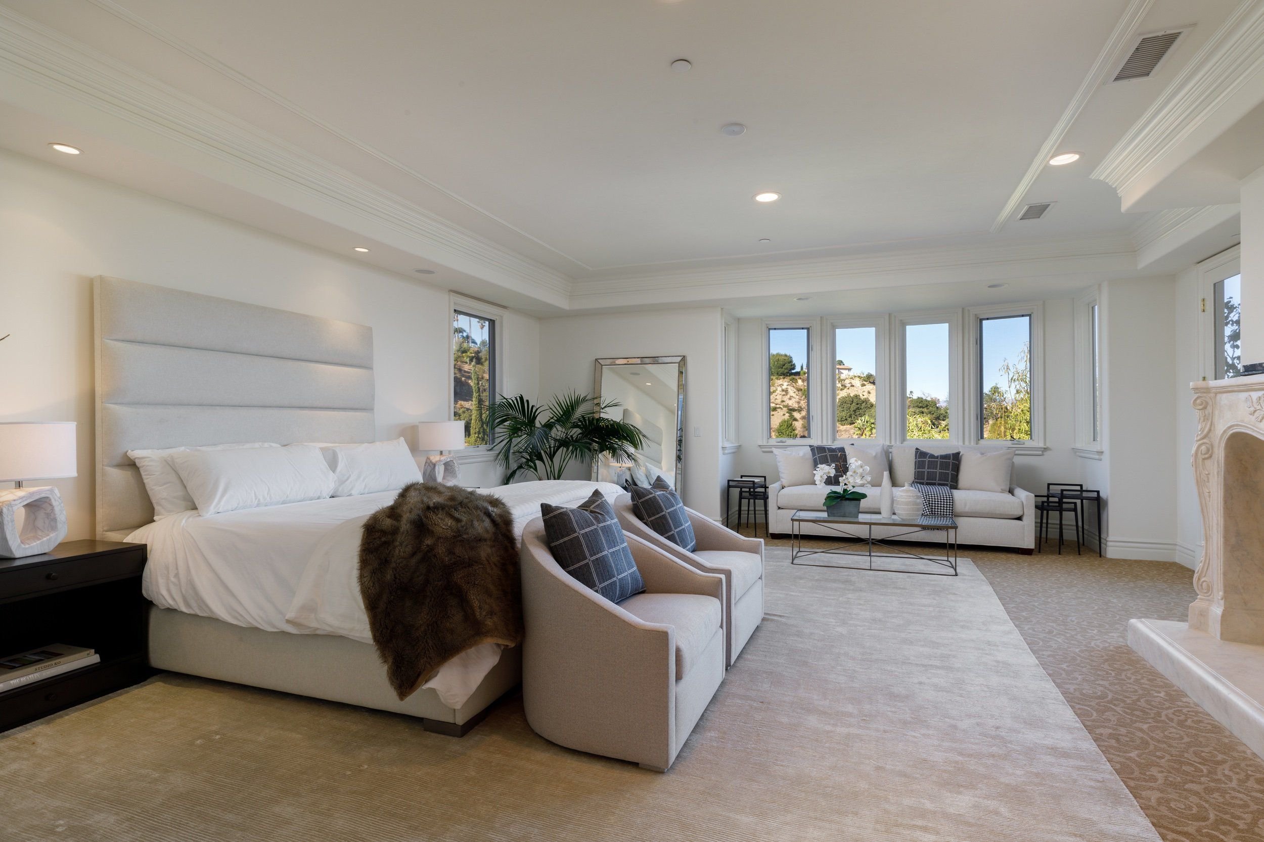 Bright bedroom with large windows showing a hilly landscape, white walls, a white upholstered bed with white bedding, a black nightstand, a beige armchair with plaid pillows, a glass coffee table with white decorative items, and various plants and mi
