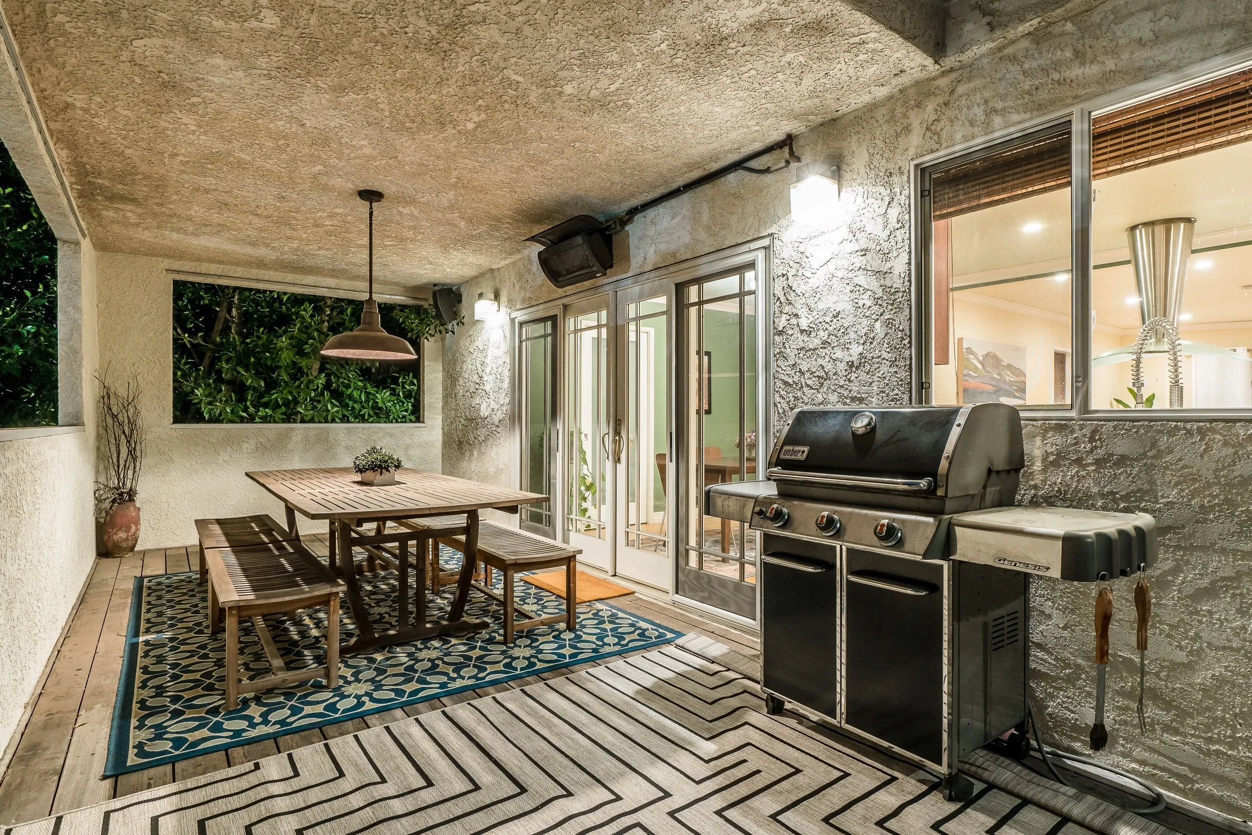 Covered outdoor patio with a wooden table, chairs, a gas grill, and decorative rugs, adjacent to glass sliding doors, with wall-mounted lights and speakers.