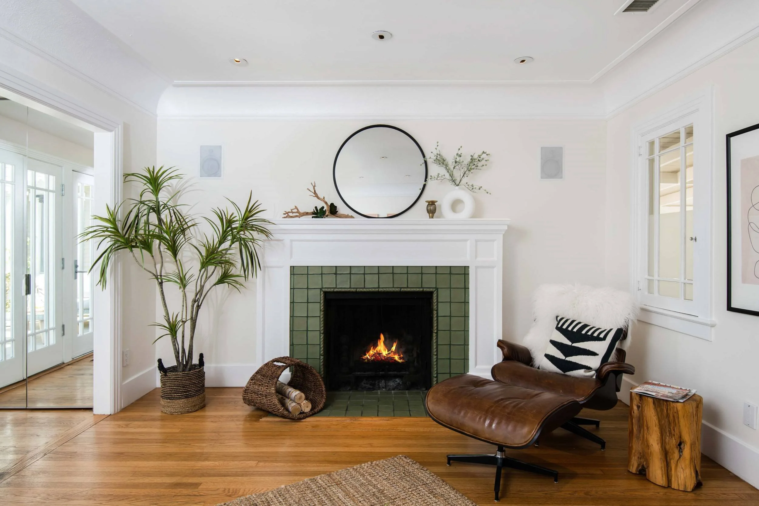 Living room with white walls, a fireplace with a fire, a round mirror above the mantel, a large green potted plant to the left, and a leather lounge chair to the right. There are decorative items on the mantel and a woven basket with logs near the fi