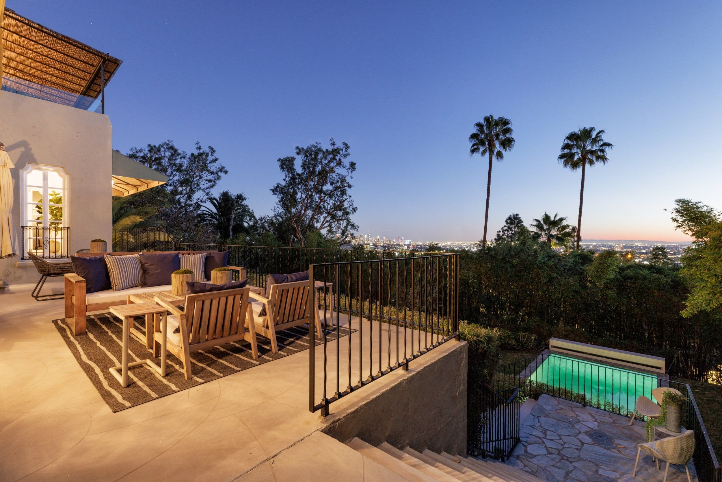 A spacious outdoor patio at dusk with comfortable seating, a striped rug, and a view of city lights in the distance, complemented by tall palm trees and lush greenery.
