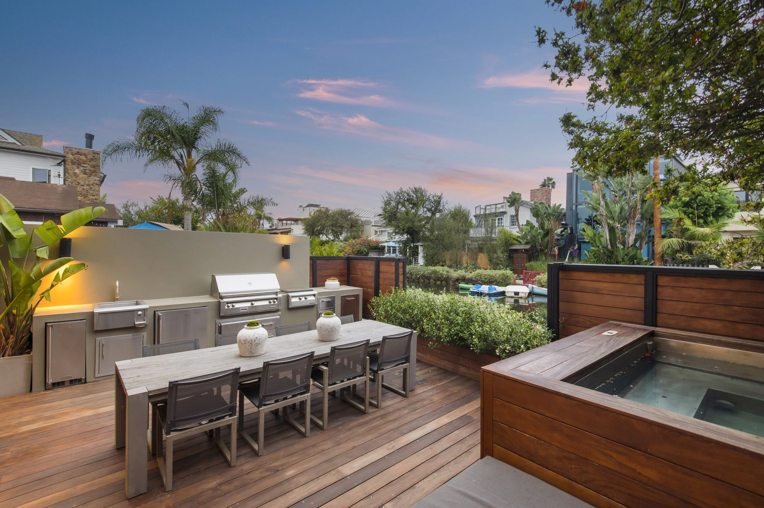An outdoor patio area with a dining table, chairs, a built-in grill, and a hot tub, overlooking neighboring houses and trees at sunset.