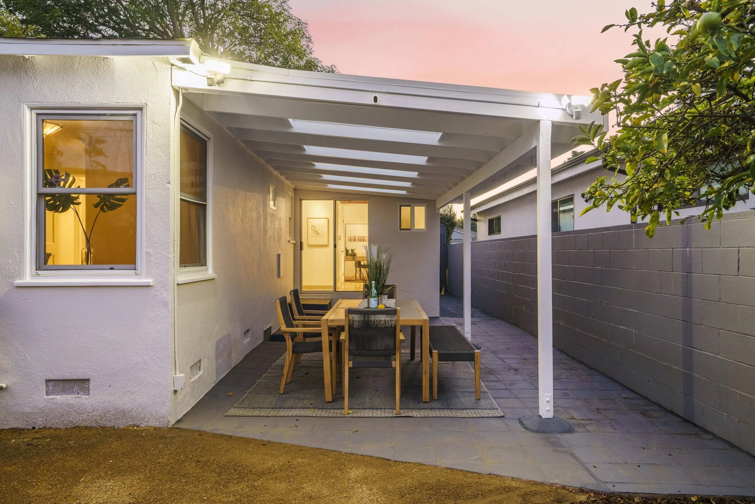A backyard patio with a dining table and chairs under a white pergola, with a sunset sky in the background. The patio is adjacent to a white house with windows, and there is a high gray wall on the right side. Greenery and tree branches are seen on t