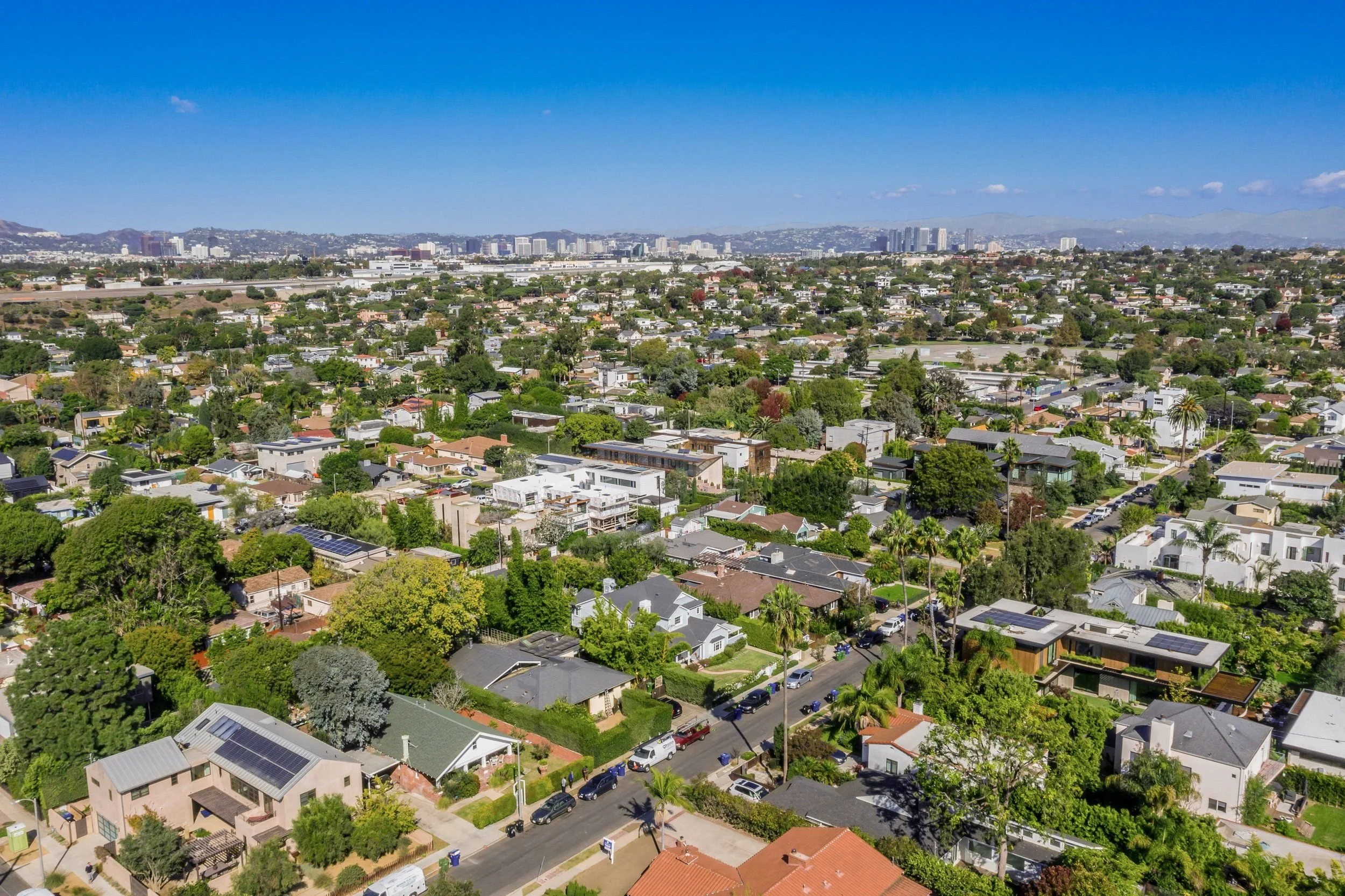 Aerial view of a suburban neighborhood with single-family homes, trees, and streets, with a cityscape and mountains in the background under a clear blue sky.