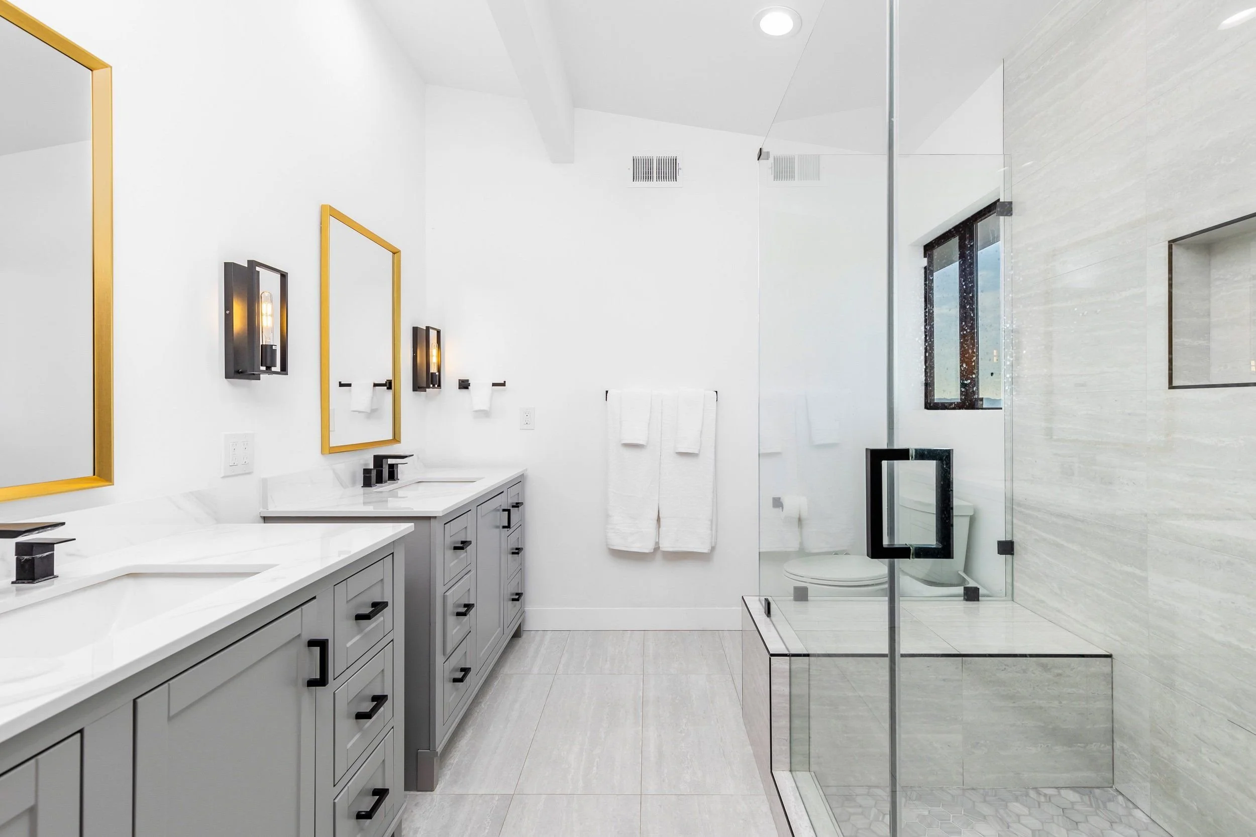 Modern bathroom with white walls, gray cabinets, black hardware, and a glass shower enclosure with marble walls and floor.