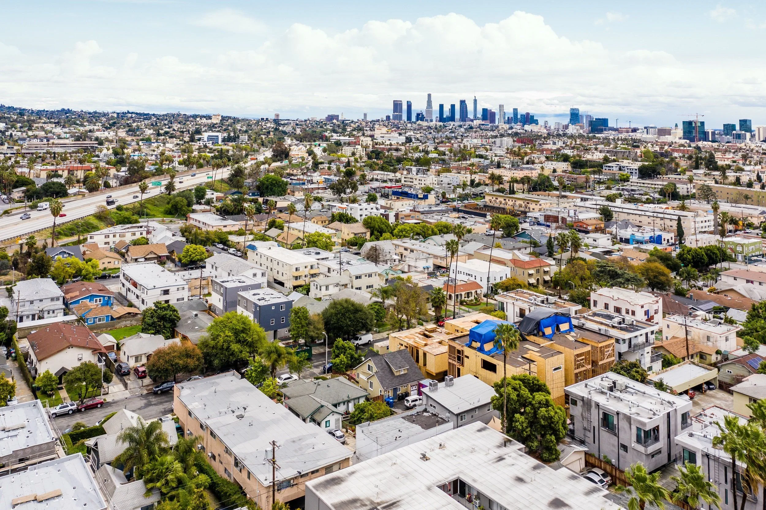 Aerial view of a city with numerous residential houses in the foreground and taller skyscrapers in the distance, under a partly cloudy sky.