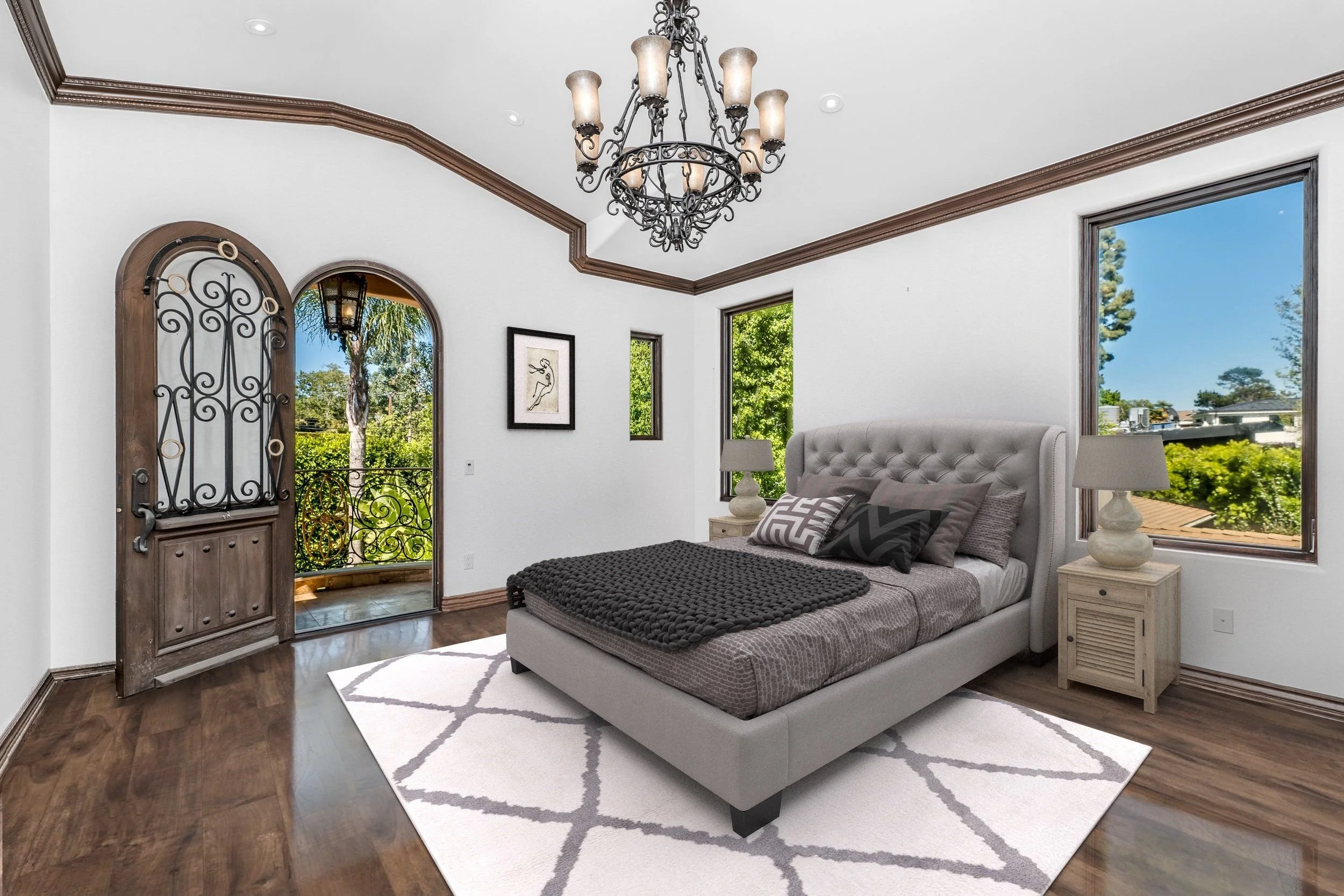 Bedroom with a grey upholstered bed, two matching nightstands with lamps, large windows showing trees outside, a decorative iron chandelier and a framed abstract art piece on the wall, white walls, hardwood floors, and a patterned rug.