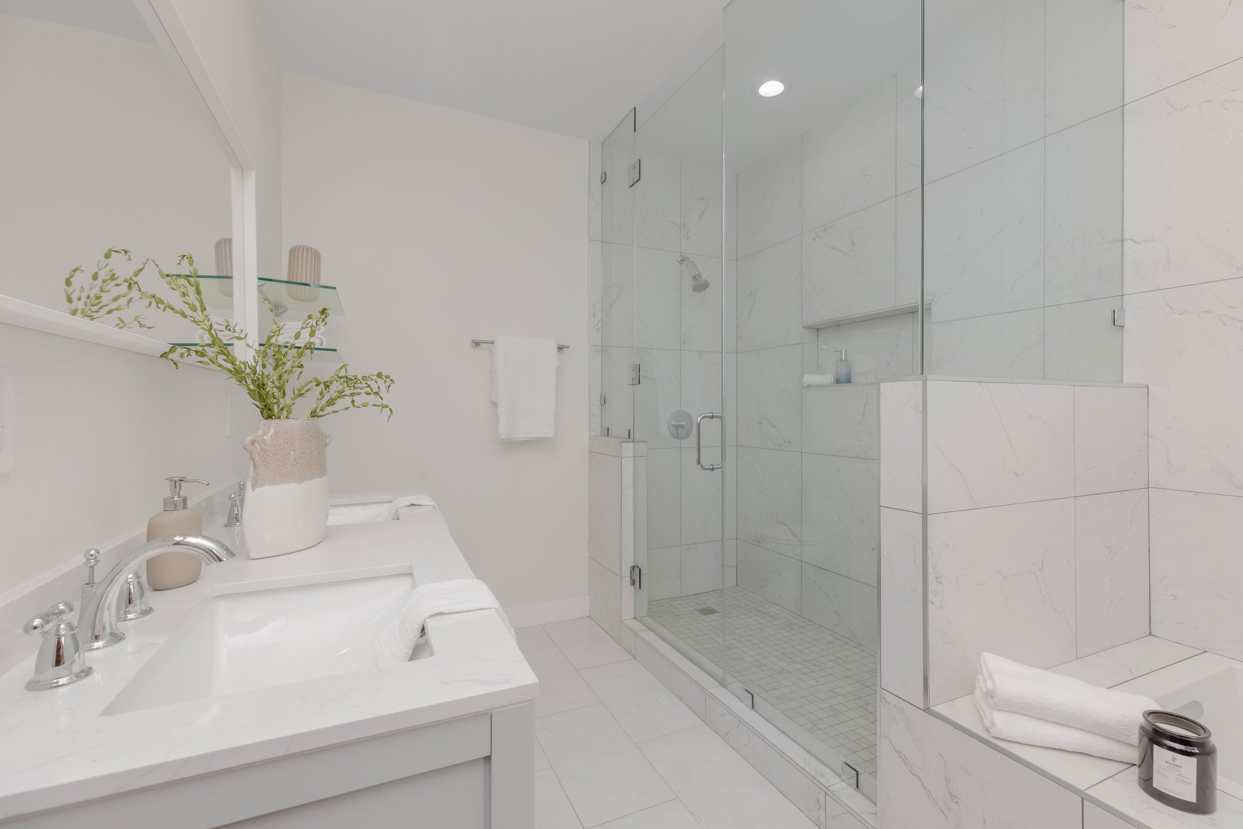 A modern bathroom with a white sink and faucet, a decorative vase with green plants, a towel on a rack, a glass-enclosed shower with marble tiles, a recessed shelf with soap, a white towel on a bench, and a dark container and candle on a nearby shelf