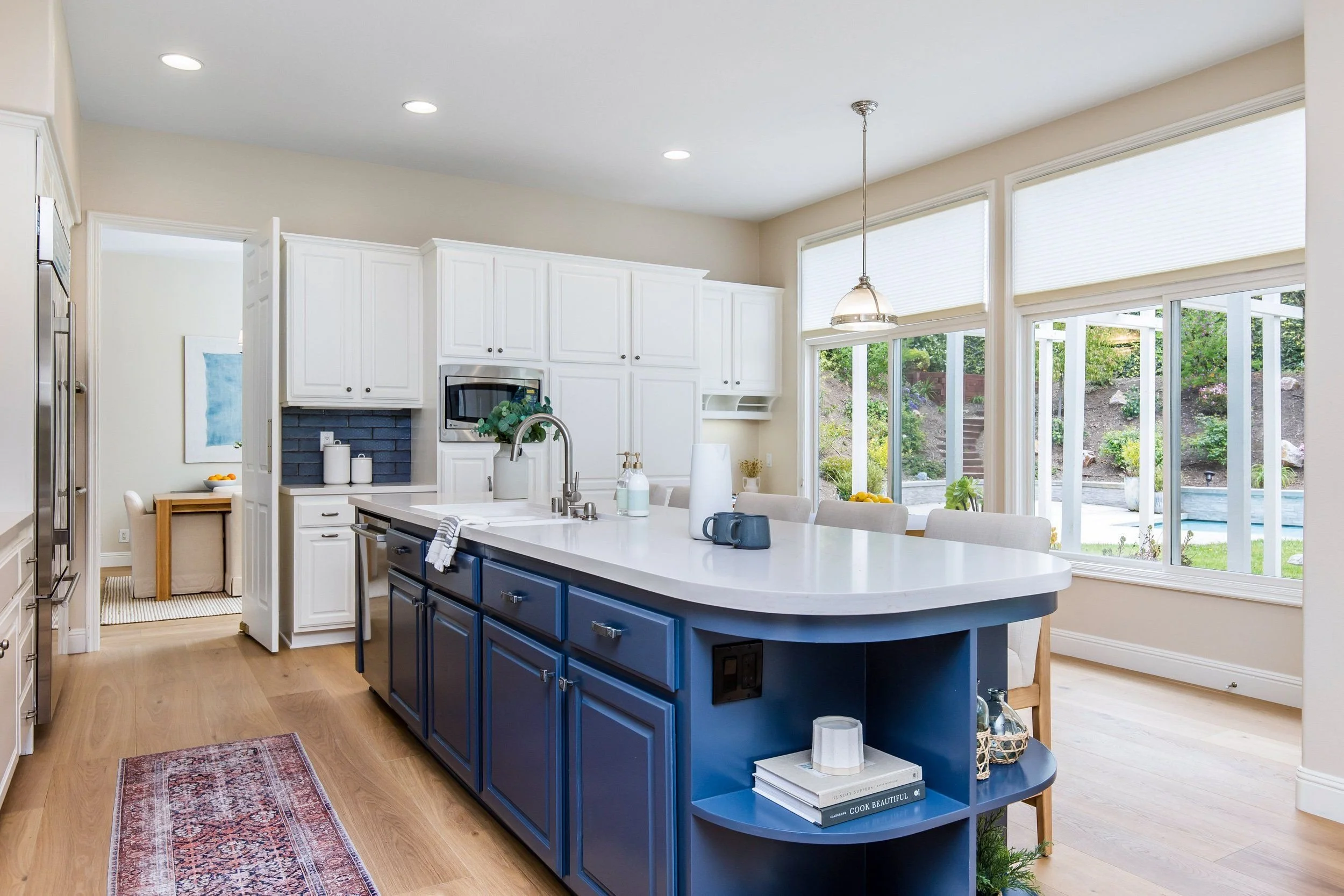 Modern kitchen with white cabinets, a blue island, large windows, and a view of a backyard with garden stairs.