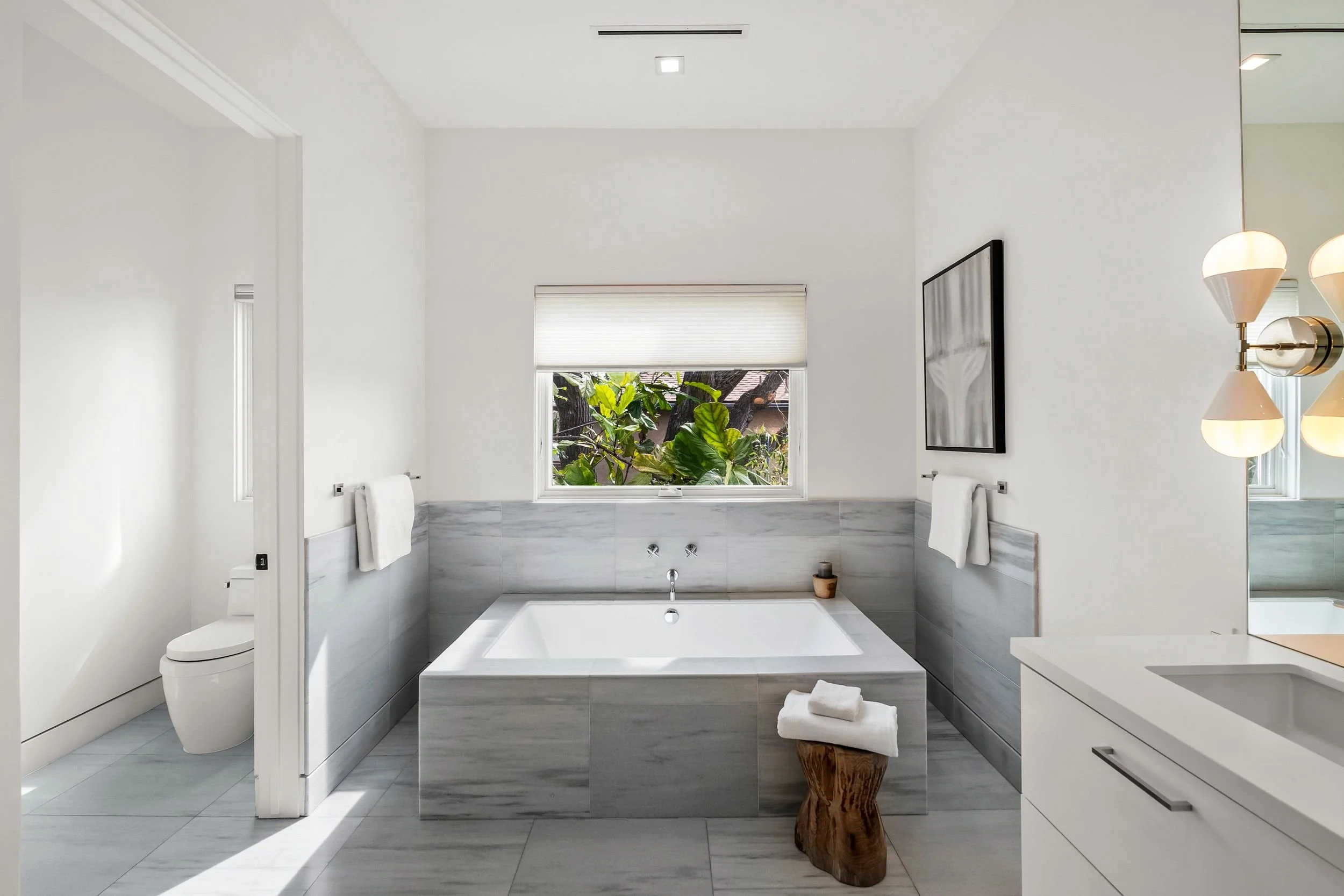 Modern bathroom with a large bathtub surrounded by gray marble tiles, a window with greenery outside, a black framed artwork, a wall-mounted lamp, and white towels.