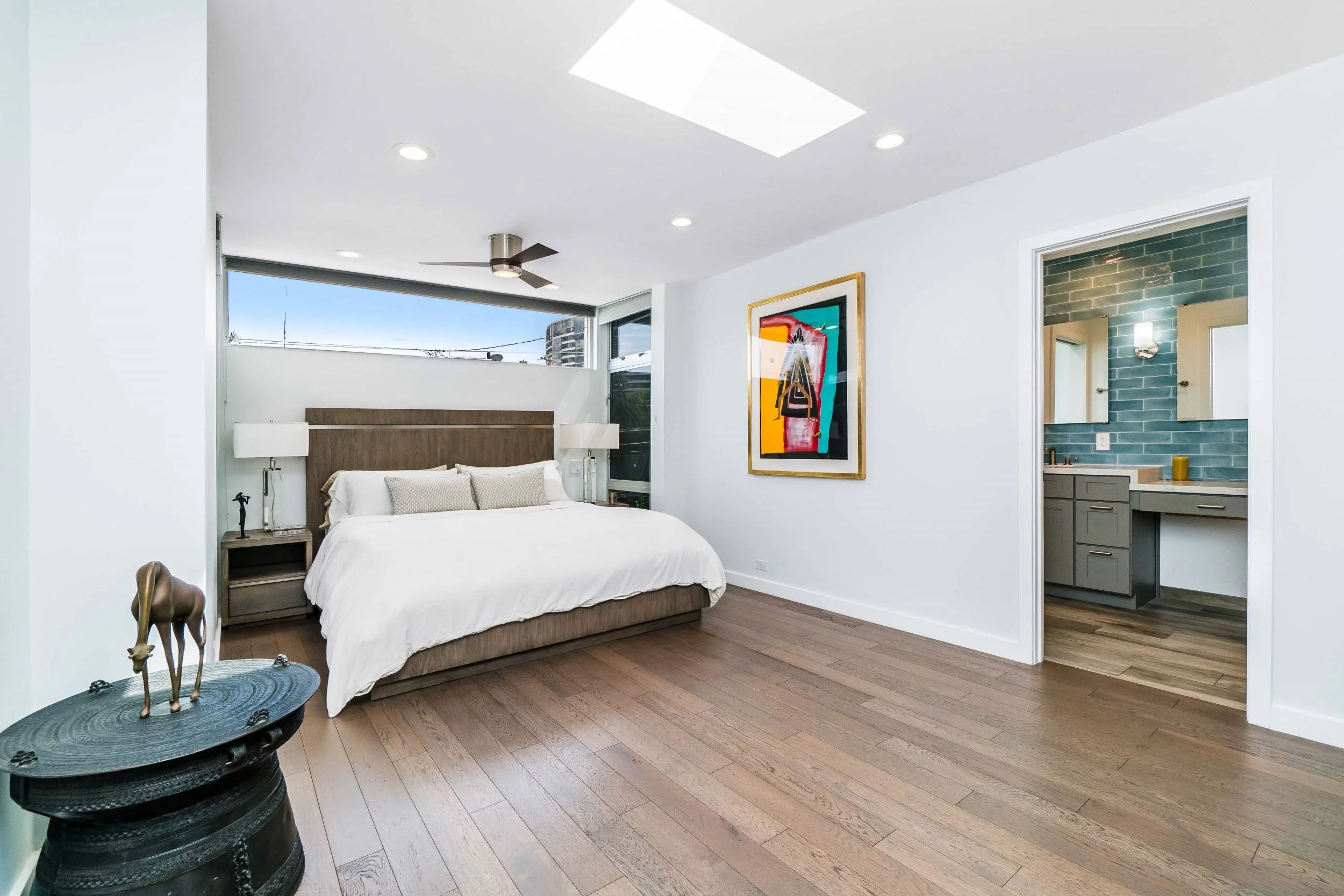 A modern bedroom with a large bed, white bedding, and two nightstands with lamps. There is a colorful abstract art piece on the wall next to a doorway leading to a bathroom with blue tiles. The room has wooden flooring and a ceiling fan.