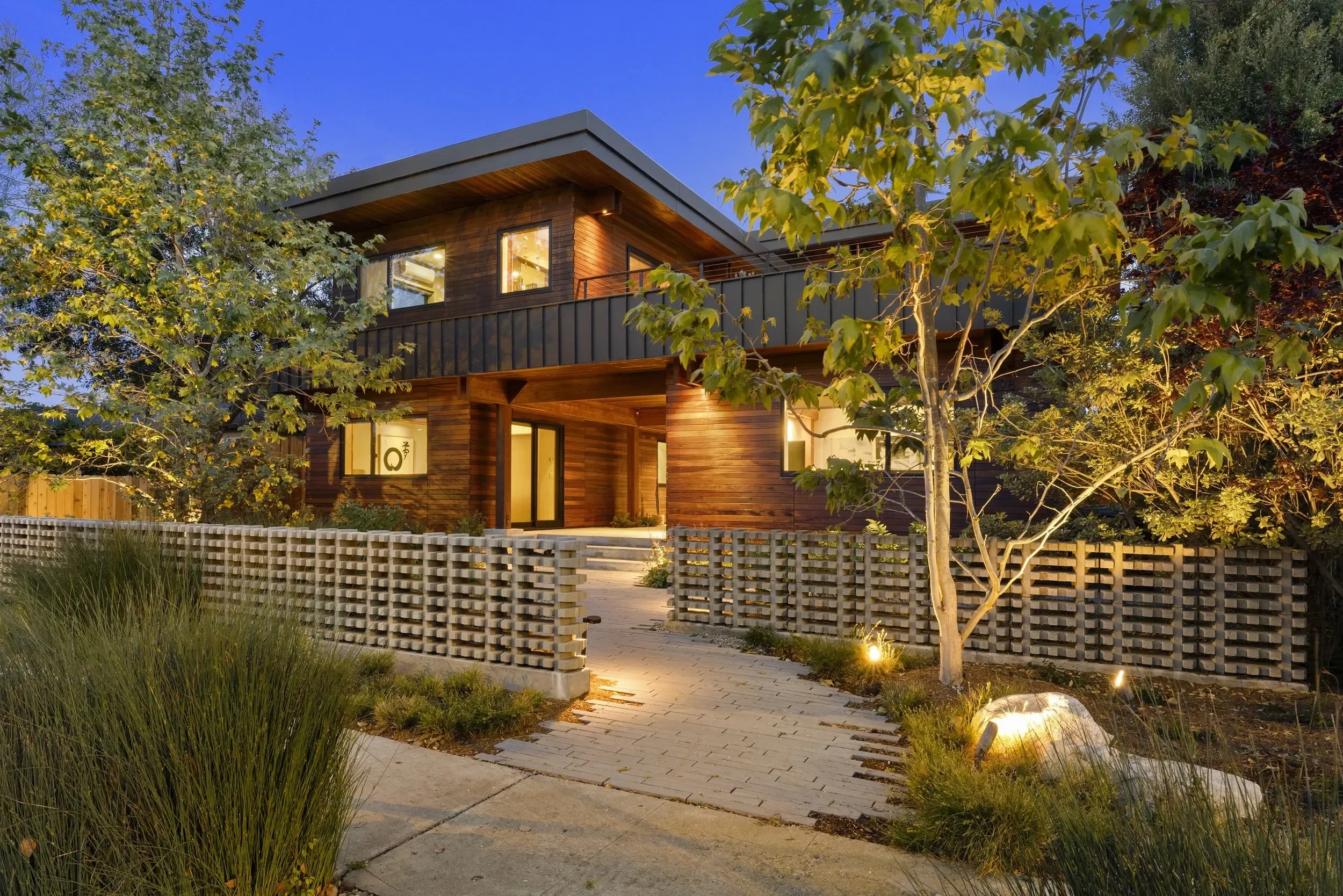 Modern two-story wooden house with large windows, surrounded by trees and landscaped garden with a brick pathway and decorative fence.
