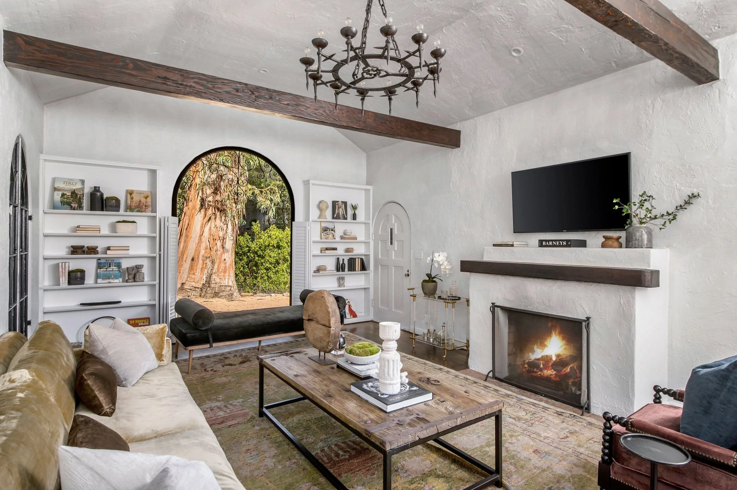 Living room with white textured walls, wooden beams, a black chandelier, and a fireplace. There are white shelves with books and decor, a large flat-screen TV, and a large window showing an outdoor scene with a tree and greenery.
