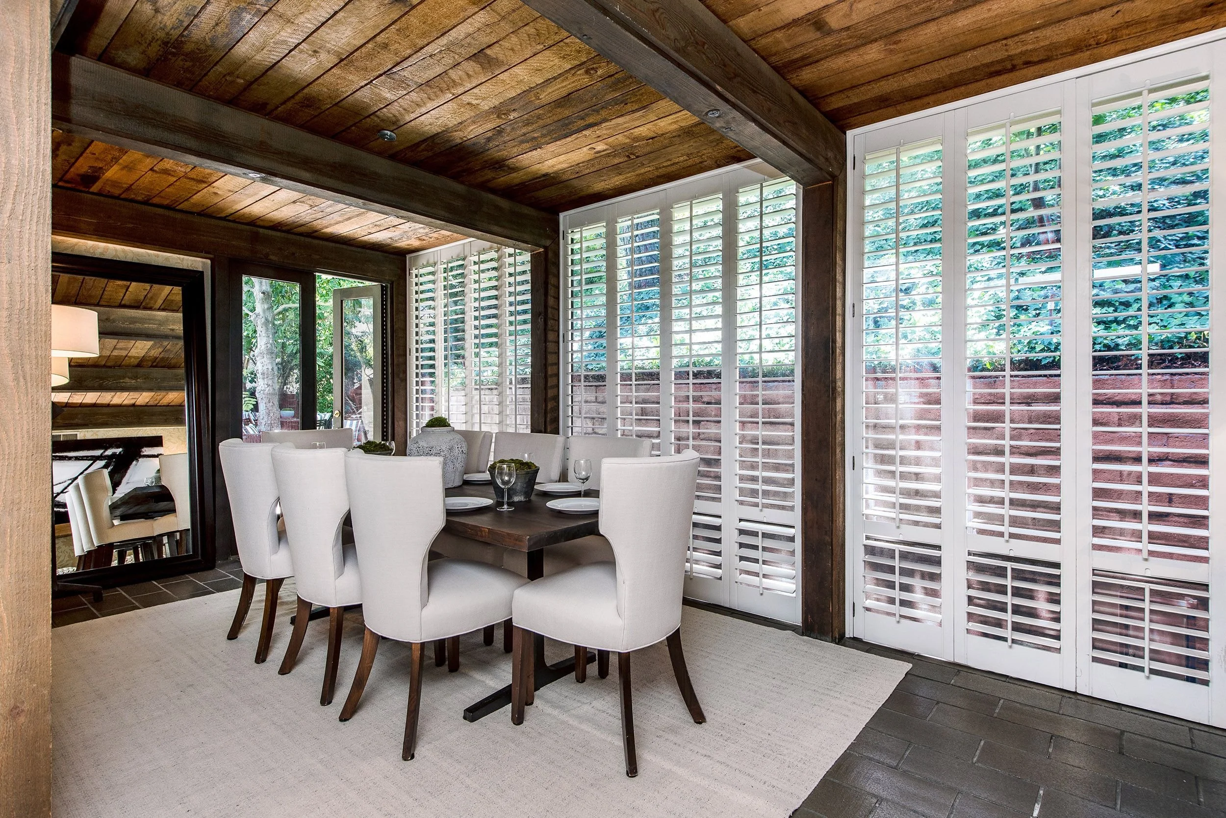 A dining room with large windows with white shutters, a wooden ceiling, and a dark wooden table surrounded by white upholstered chairs. There are place settings and decorative vases on the table.