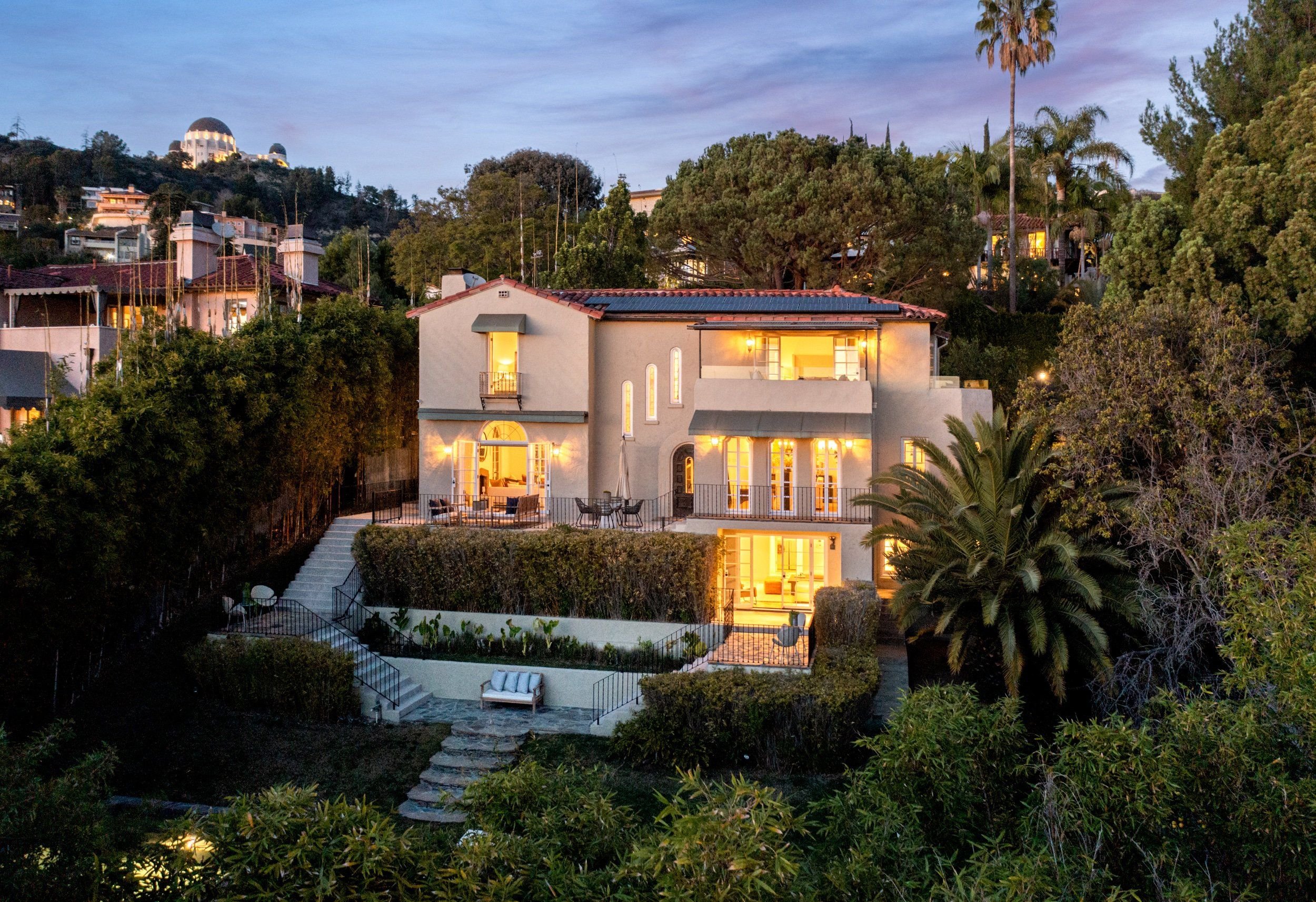 A multi-story house with warm interior lighting, surrounded by trees and shrubs, located in a hillside suburban area during evening.