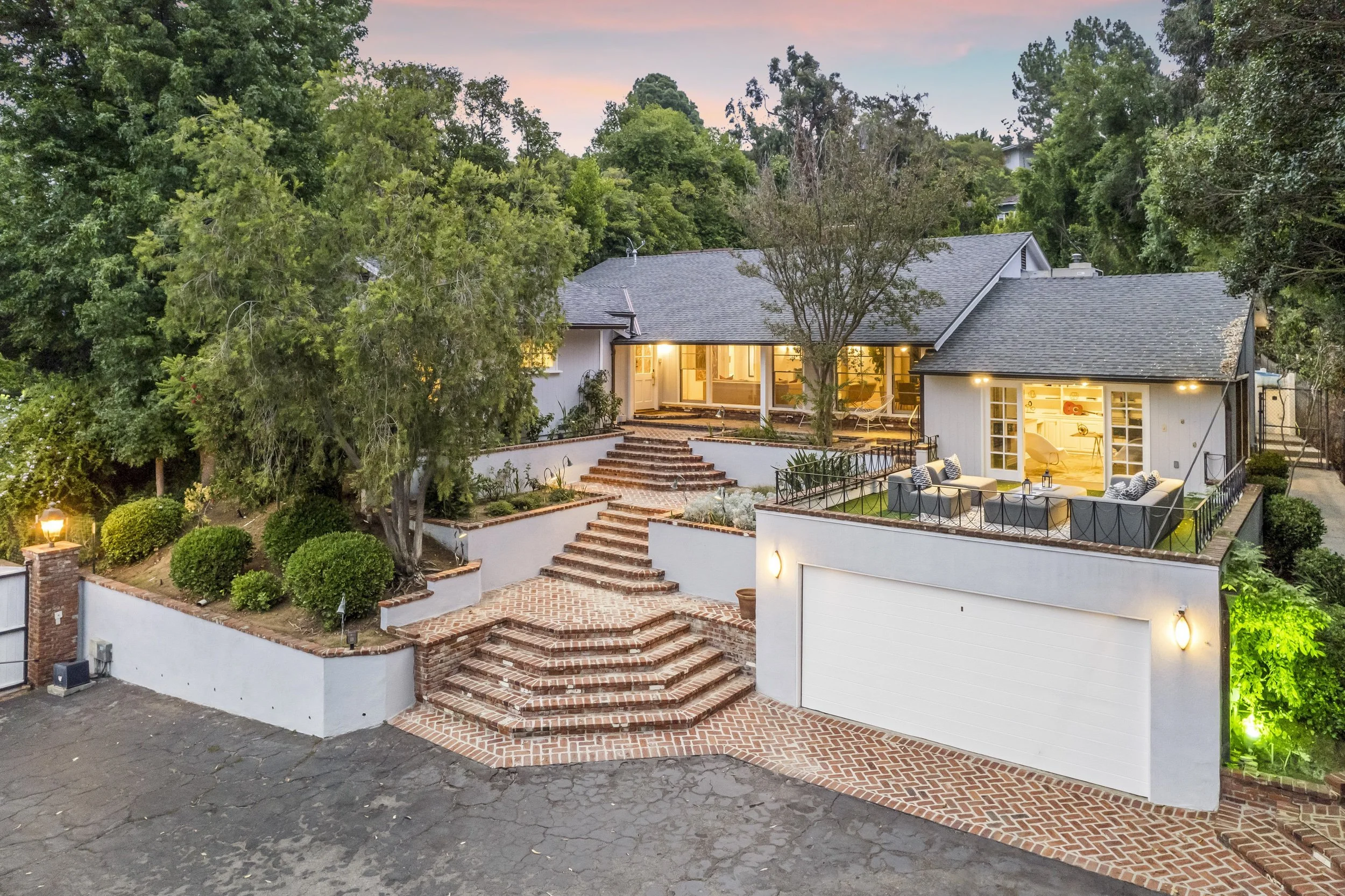A house with a large backyard, illuminated outdoor living space, brick stairs leading up to it, surrounded by trees and bushes, with a sunset sky in the background.