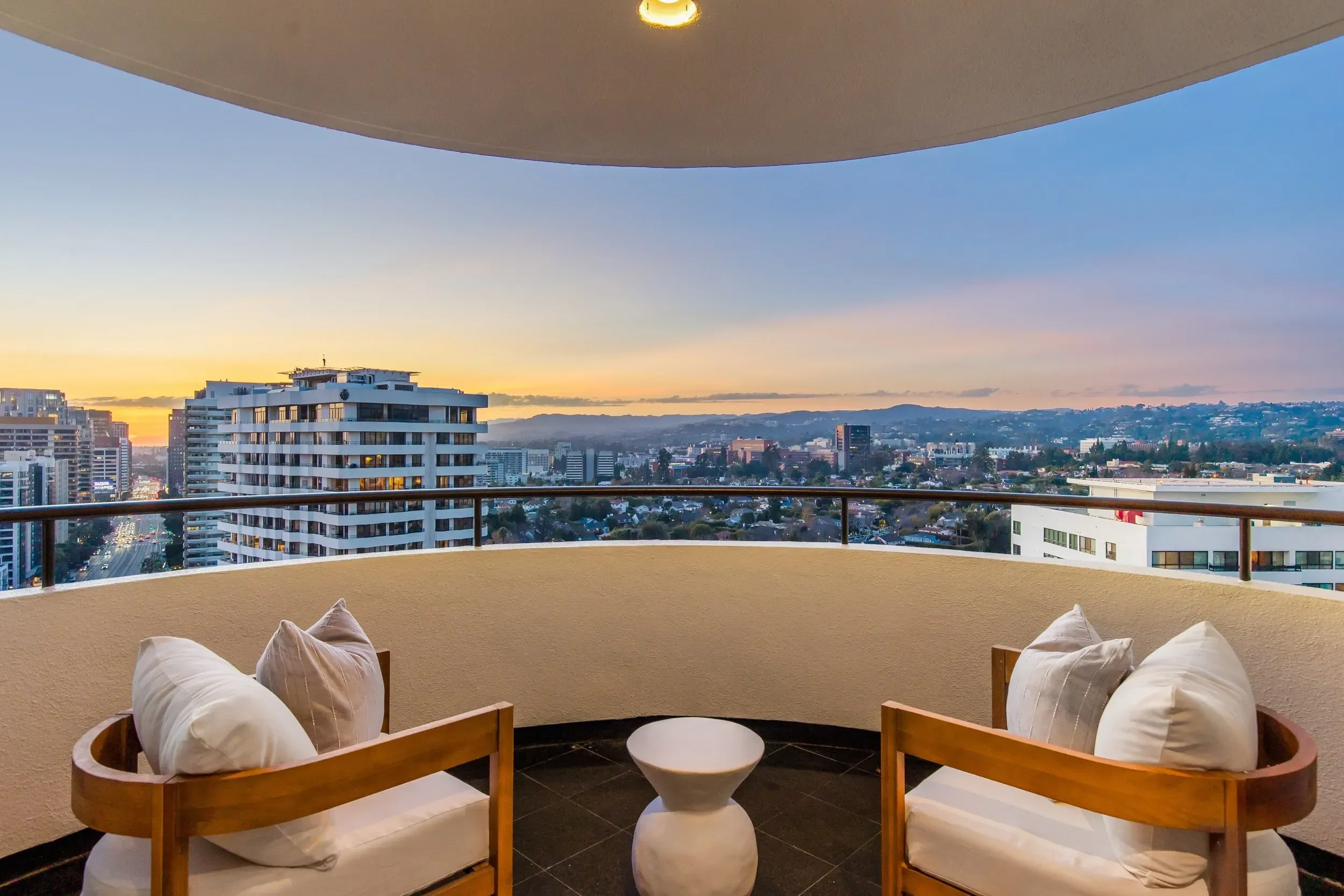 A balcony with four cushioned chairs arranged in a semi-circle around a small round table. The balcony offers a panoramic view of a city skyline at sunset, with various tall buildings and hills in the distance, under a colorful sky.