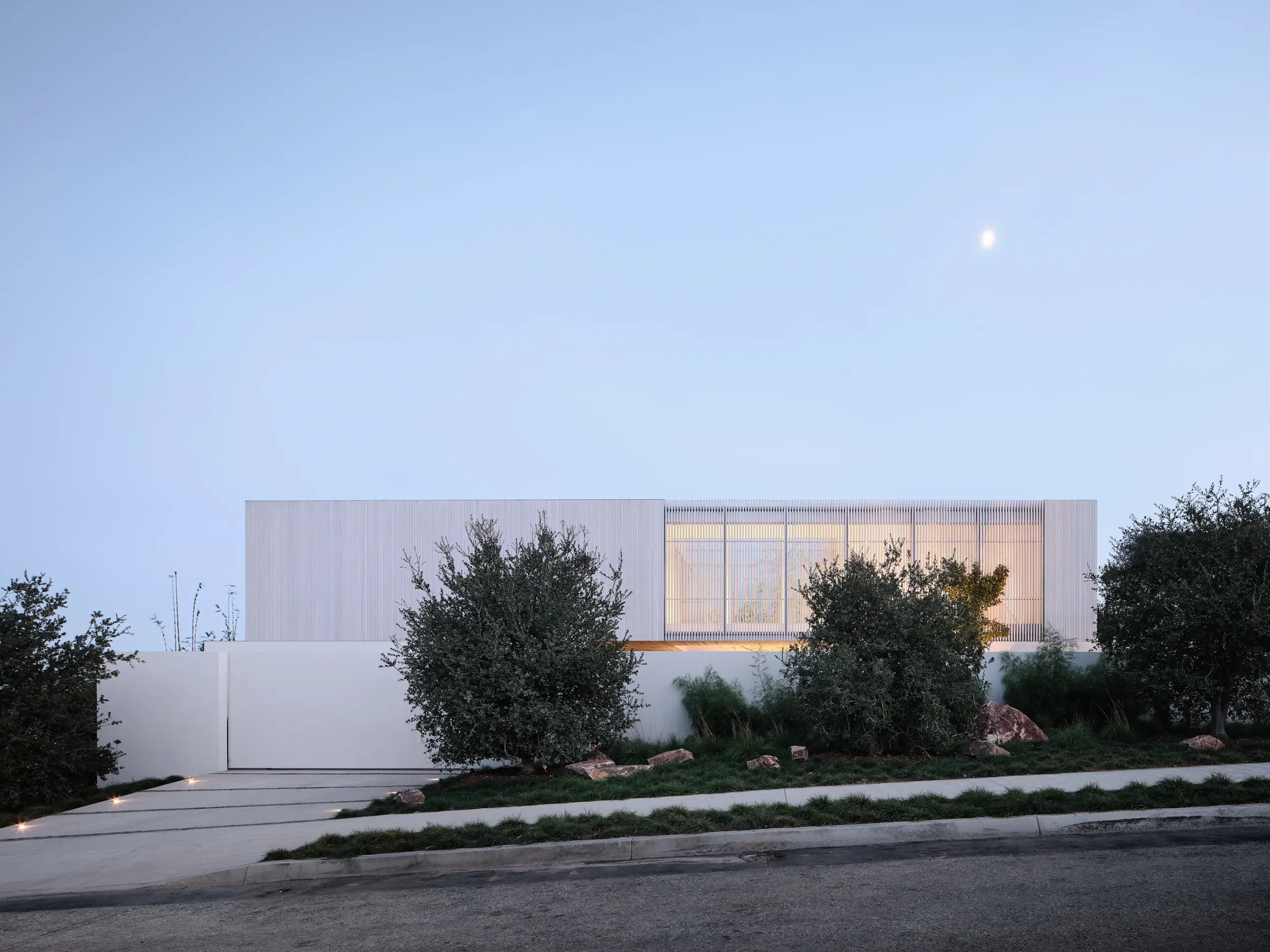 Modern building with a perforated metal facade illuminated from inside, surrounded by trees and plants, under a clear evening sky with visible moon.