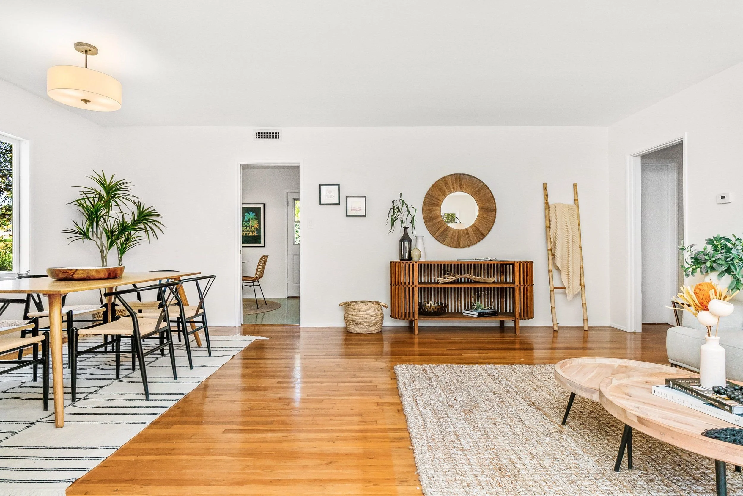 Bright, modern living room with hardwood floors, white walls, plants, a wooden sideboard with vases, and wall art.