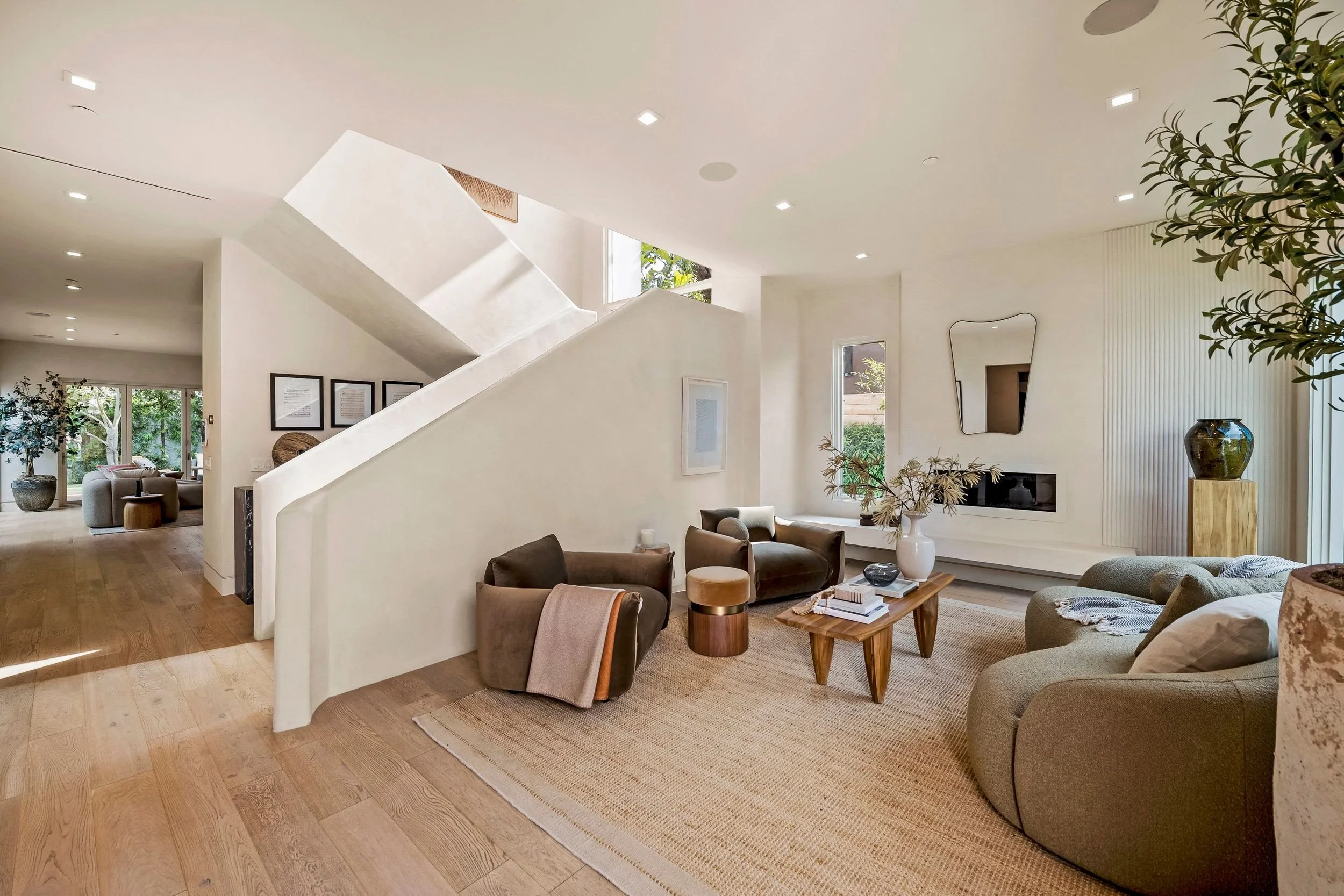 Modern living room with beige sofas, a wooden coffee table, and potted plants, featuring high ceilings, large windows, and a staircase.